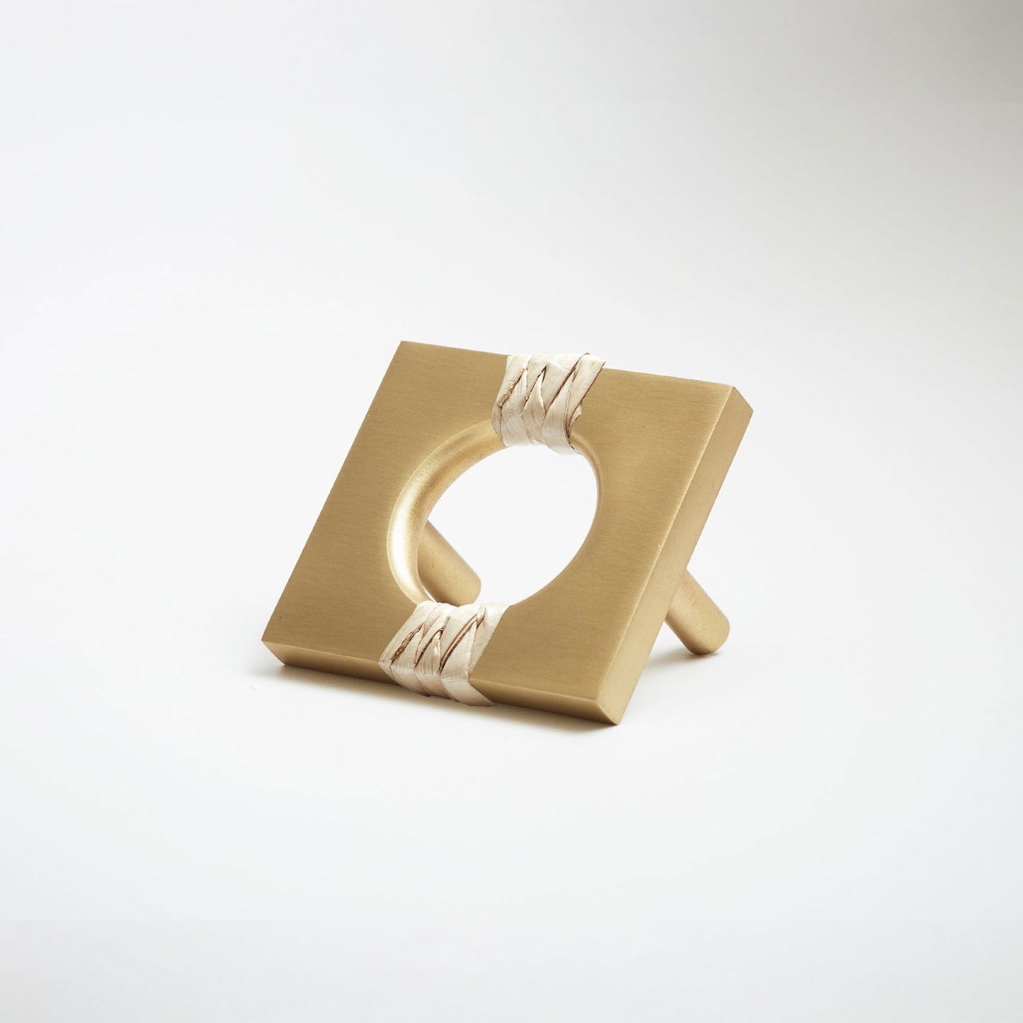 A satin brass metal geometric cabinet pull with a square frame and circular center, wrapped with a light natural leather at the corners, shown on a white background.