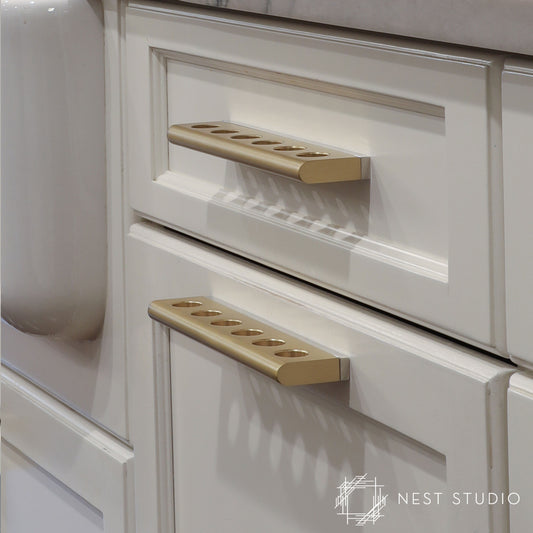 Close-up of a cream-colored kitchen cabinet with two drawers, each fitted with a modern rectangular pull handle in a warm brushed gold finish. The cabinet features raised panel detailing and a light stone countertop is partially visible at the top.