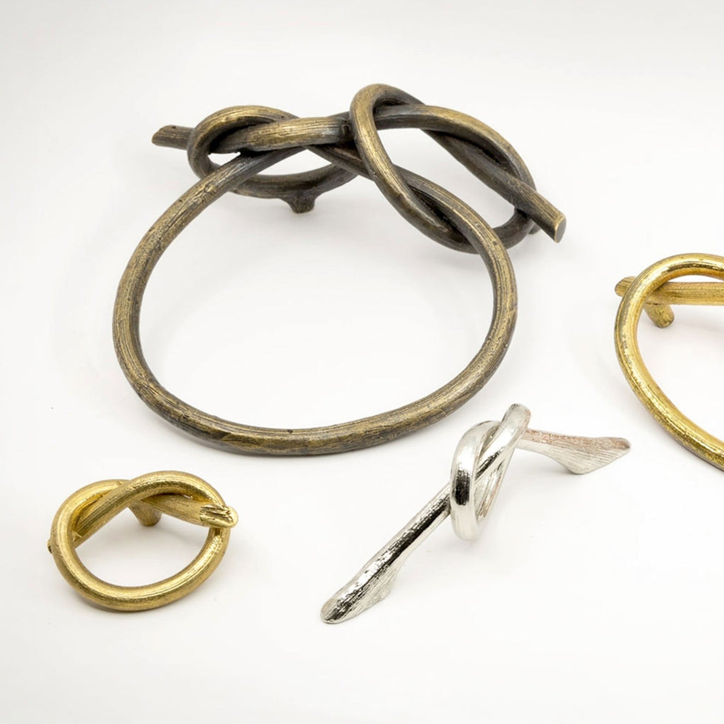 Three sculptural knot-style cabinet pulls displayed on a clean white background: one large antiqued bronze piece, one small gold-toned piece, and one small silver-toned piece.