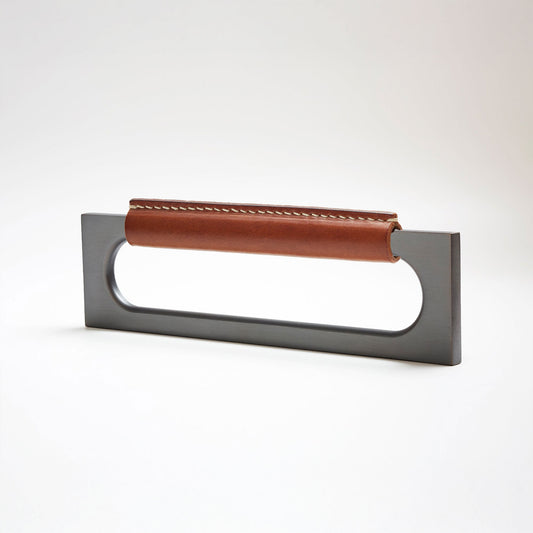 A modern rectangular metal cabinet pull with a central cutout, featuring a light brown leather strap stitched across the top, shown on a clean grey studio background.