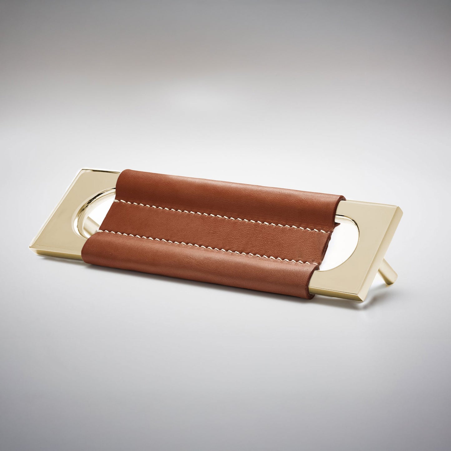 A modern rectangular metal cabinet pull with a central cutout, featuring a light brown leather strap stitched across the top, shown on a clean grey studio background.