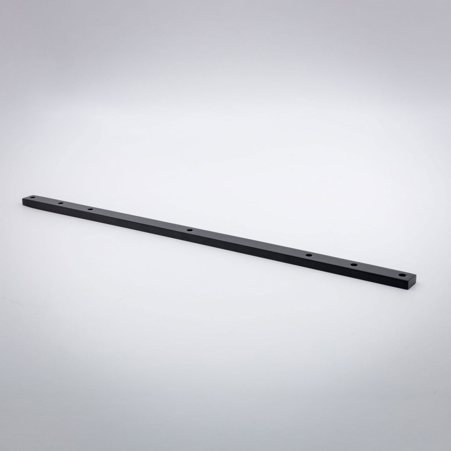 A long, thin black brass metal bar with pre-drilled holes, photographed diagonally on a light gray gradient background.