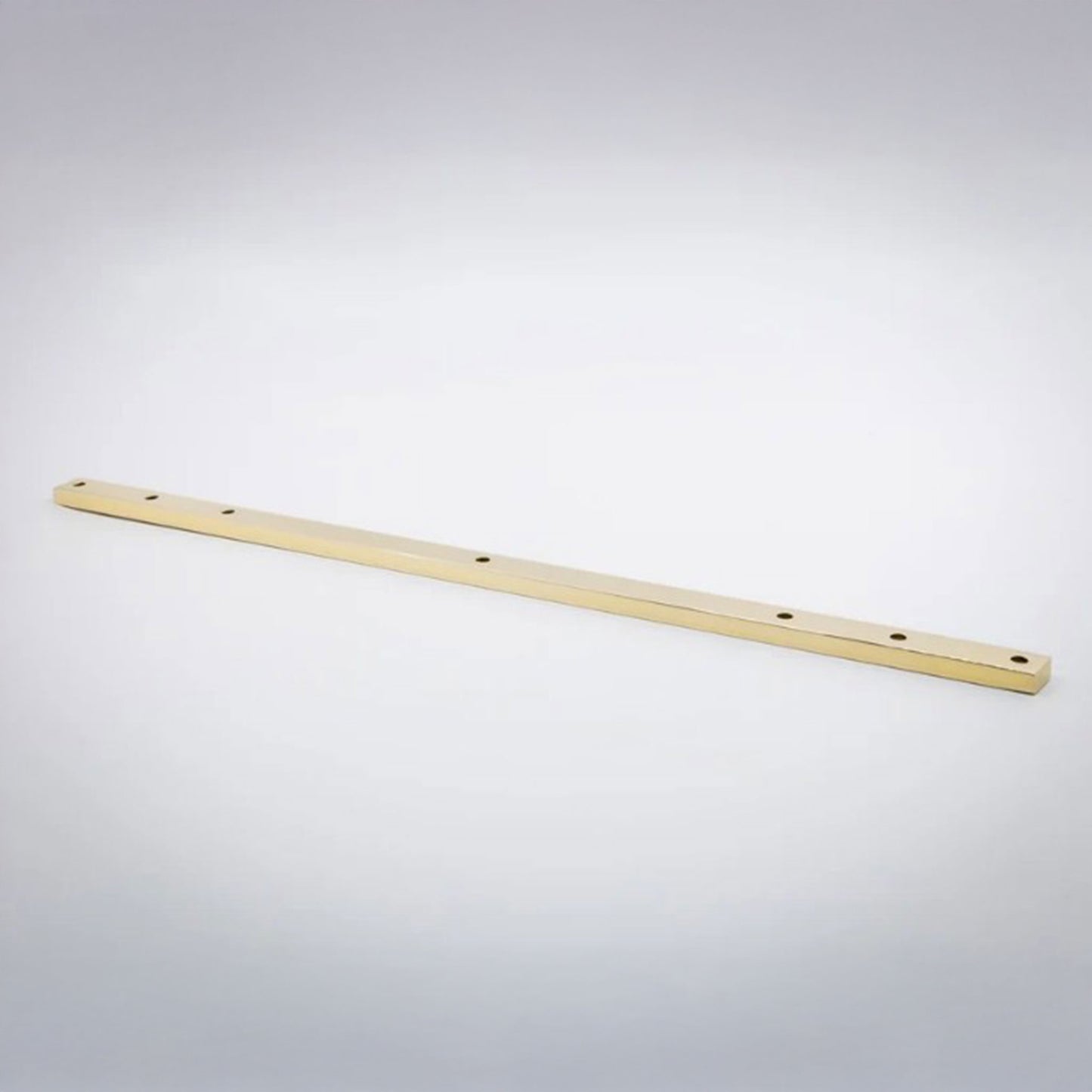 A long, thin unlaquered polished brass metal bar with pre-drilled holes, photographed diagonally on a light gray gradient background.