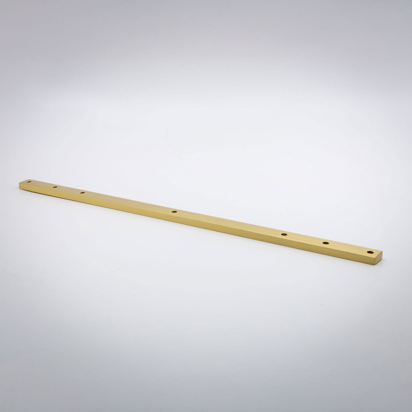 A long, thin satin brass metal bar with pre-drilled holes, photographed diagonally on a light gray gradient background.