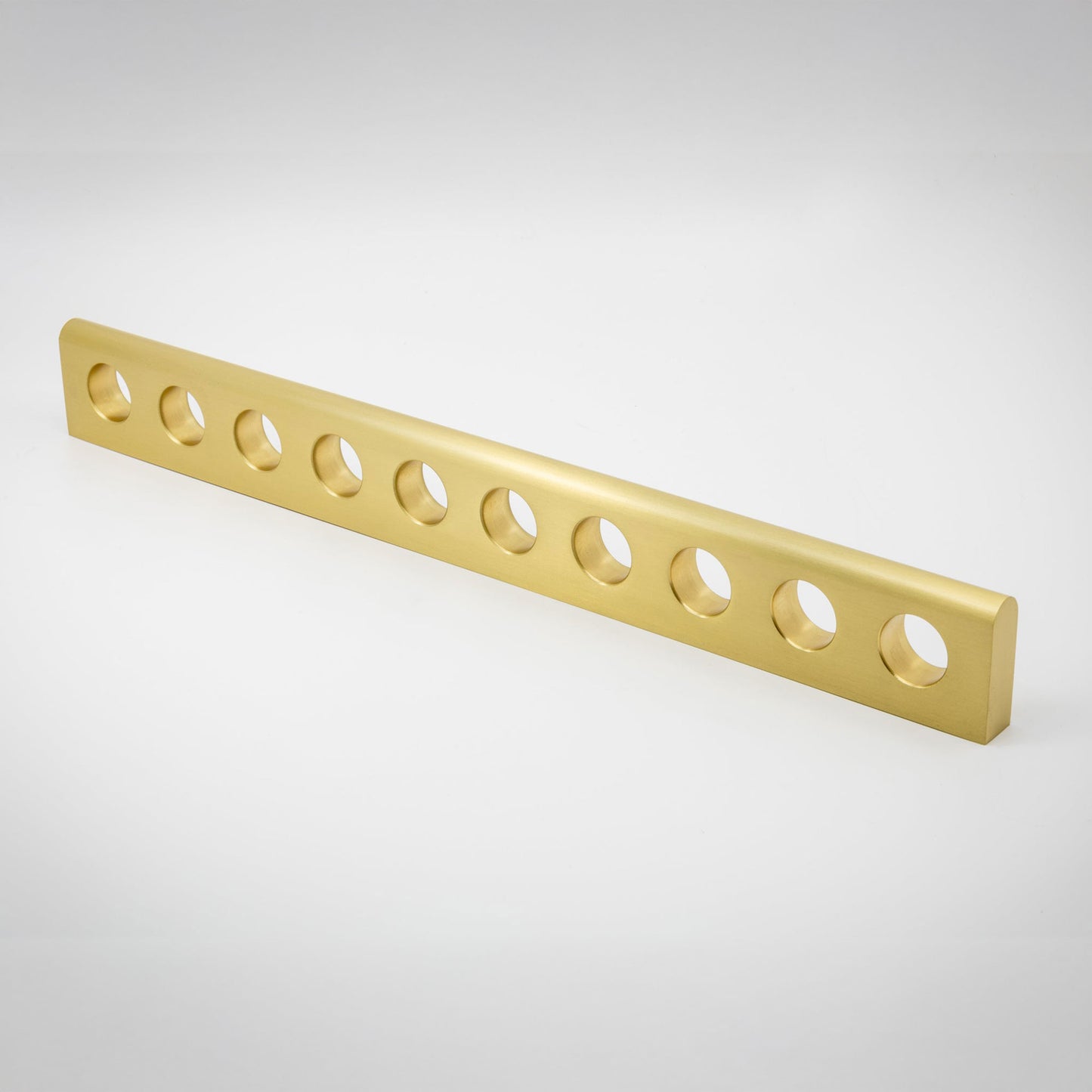 A long, narrow satin brass gold metal bar with ten evenly spaced circular cutouts, shown diagonally on a light gray gradient background.