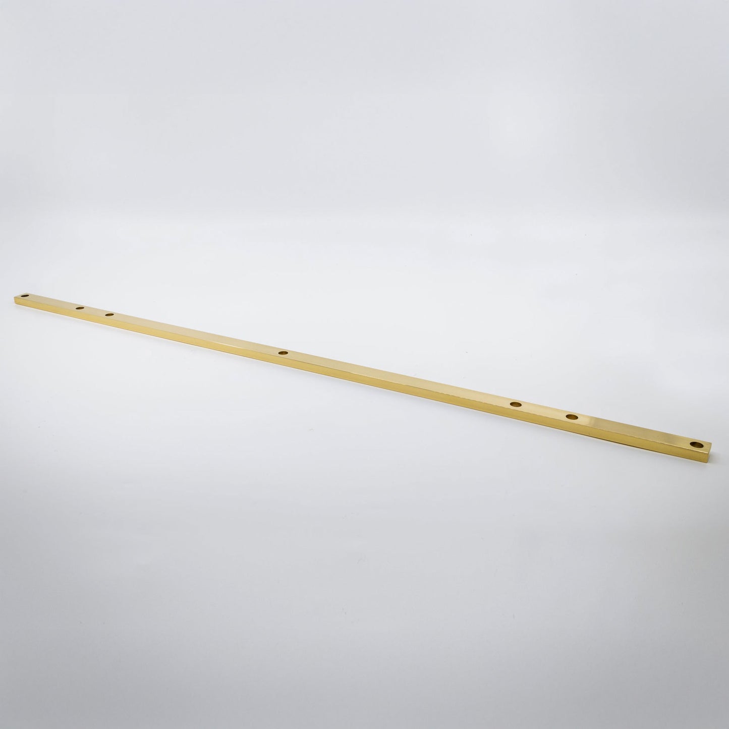 A long, thin metallic gold bar with pre-drilled holes, photographed diagonally on a seamless light gray background.