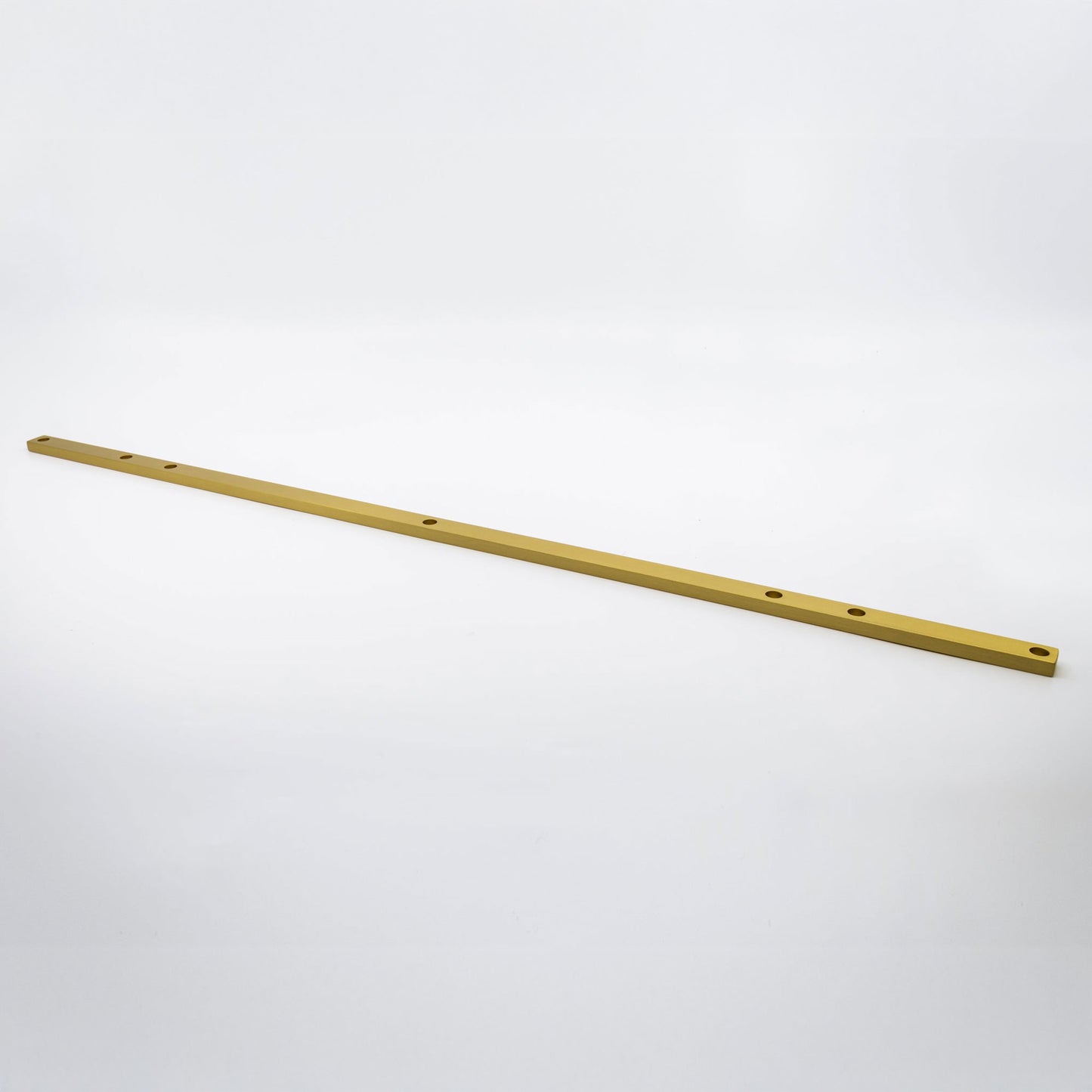 A long, thin metallic gold bar with pre-drilled holes, photographed diagonally on a seamless light gray background.