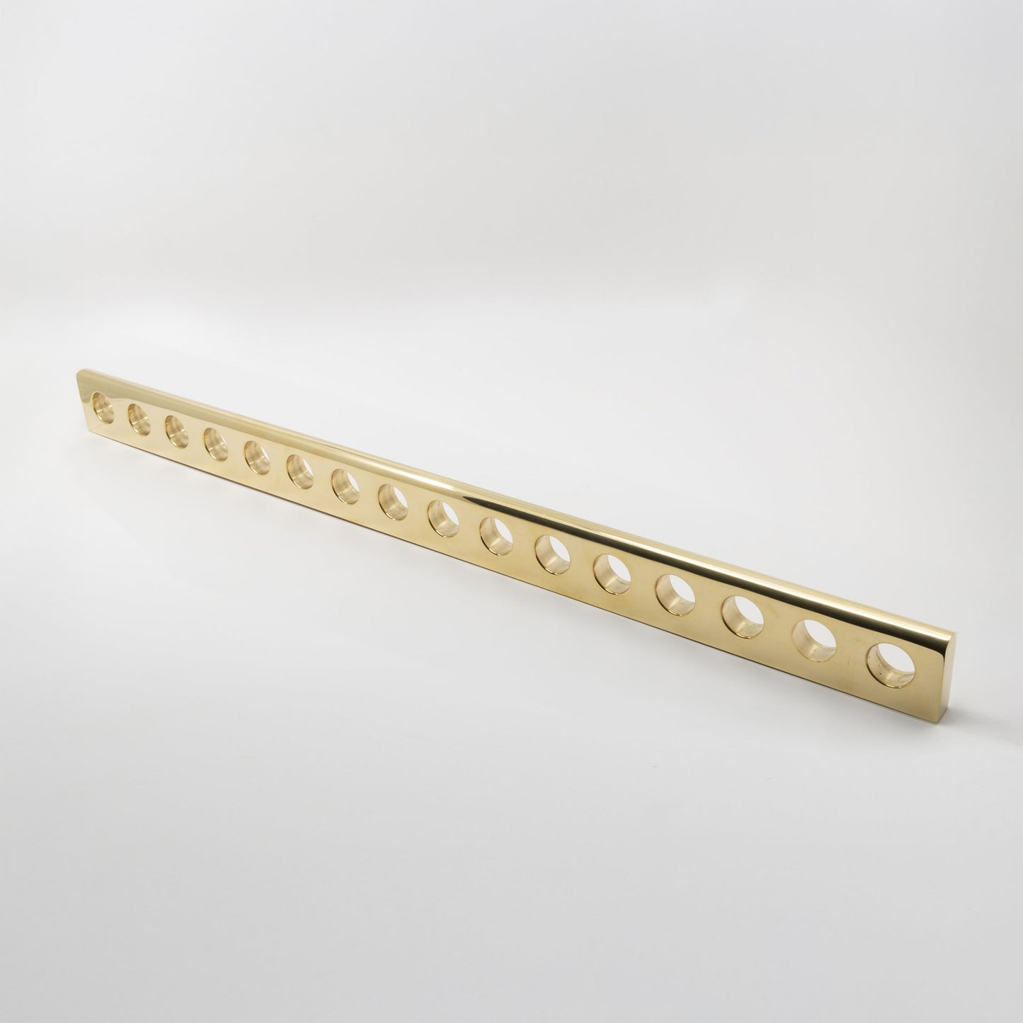 A long, slender gold unlaquered brass metal hardware handle with multiple evenly spaced circular cutouts, photographed diagonally on a clean light gray studio background.