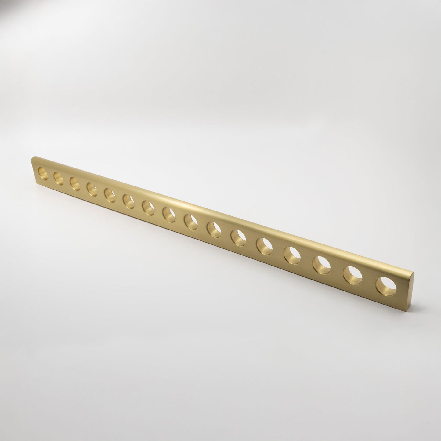 A long, narrow metallic brass bar with multiple circular cutouts, shown diagonally on a clean light gray studio background.