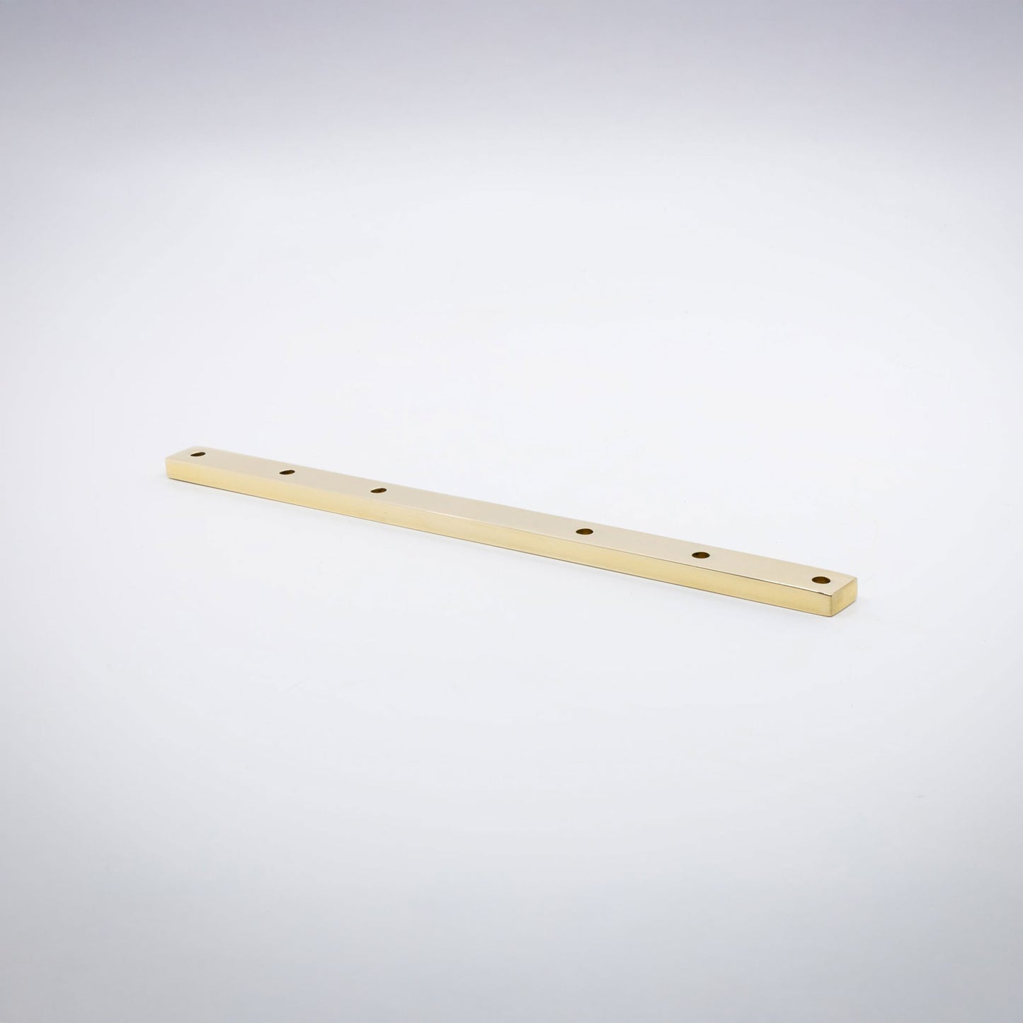 A long, narrow rectangular unlaquered polished brass base hardware bar with six evenly spaced circular holes, shown diagonally on a clean light gray studio background.