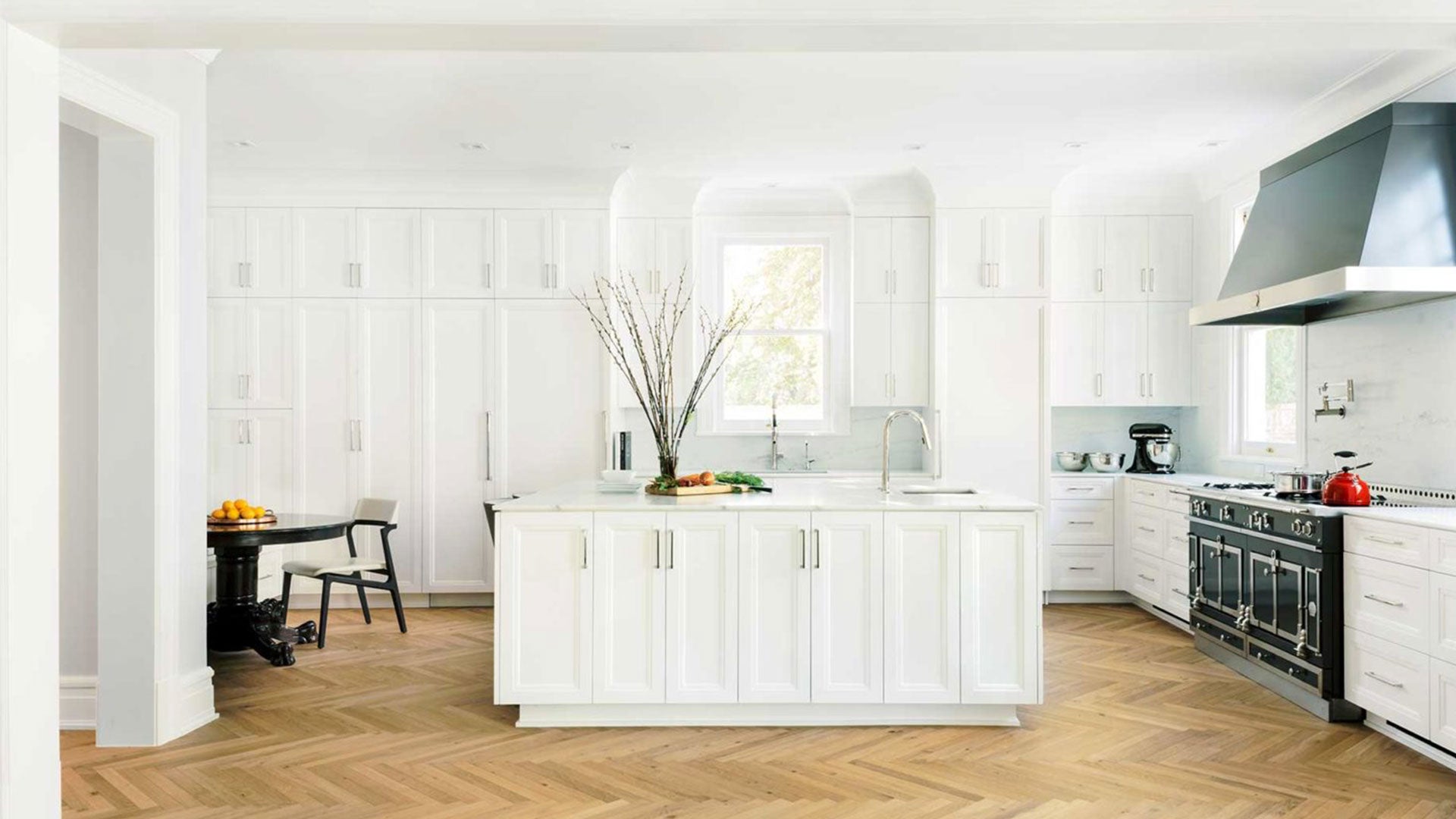 A classic white kitchen with black accents including a luxury black enamel stove and featuring a pale oak chevron wood floor.