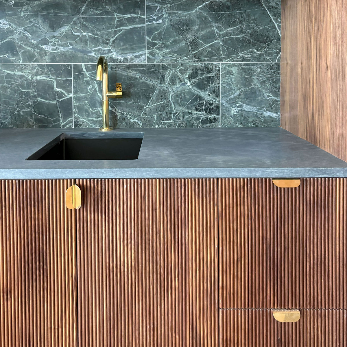A gold faucet on a stone slab surrounded by wood cabinetry and matching god tab pulls