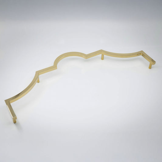 A long decorative cabinet pull handle with a wavy scalloped edge, in a polished unlacquered brass shown on a clean light gray studio background.