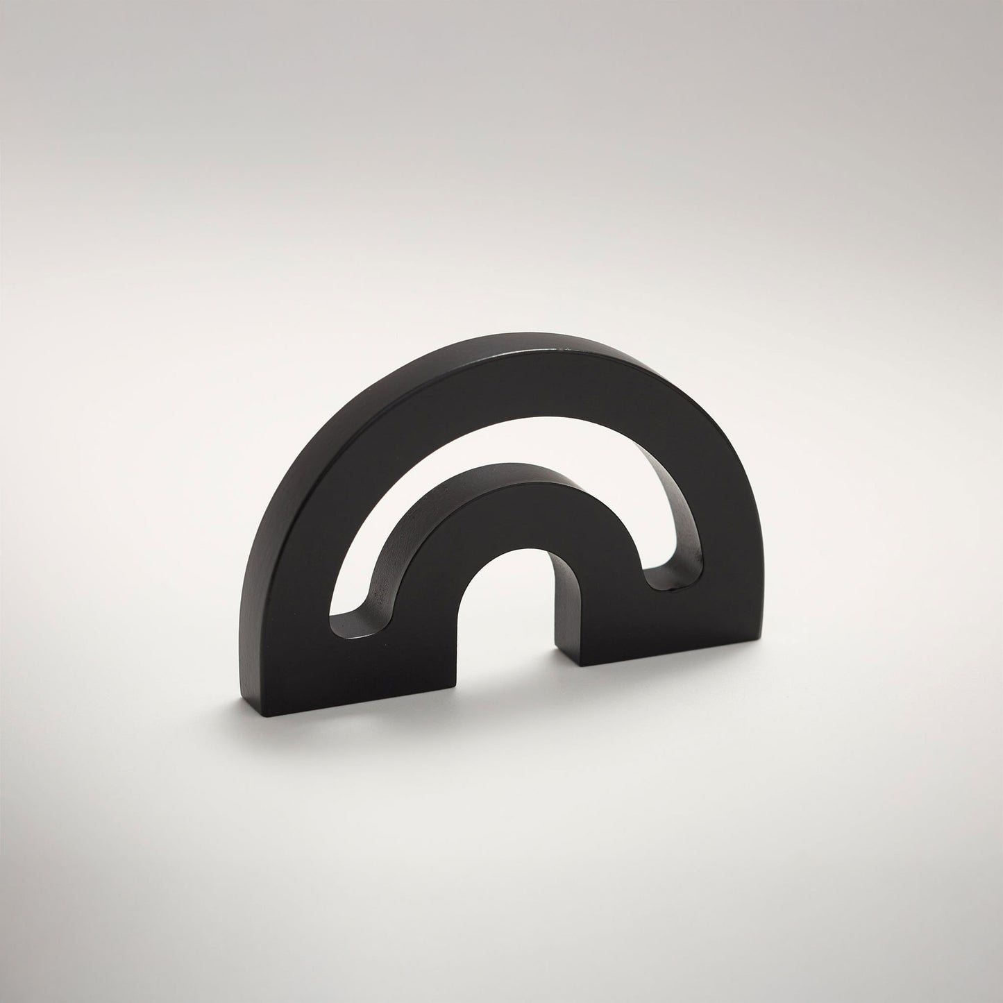 A minimalist metal cabinet knob with a double-arch design, photographed on a clean light gray gradient background.