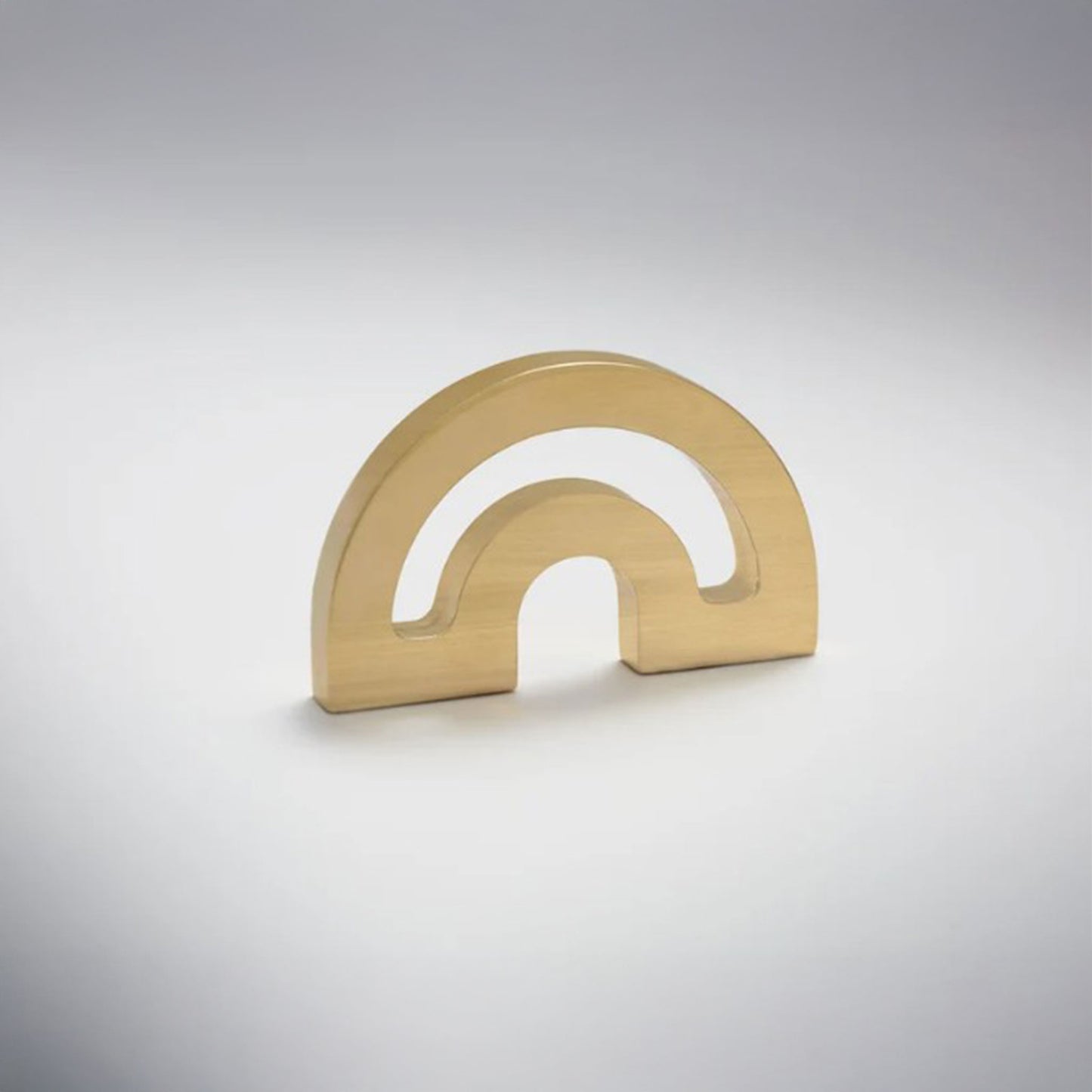 A minimalist metal cabinet knob with a double-arch design, photographed on a clean light gray gradient background.