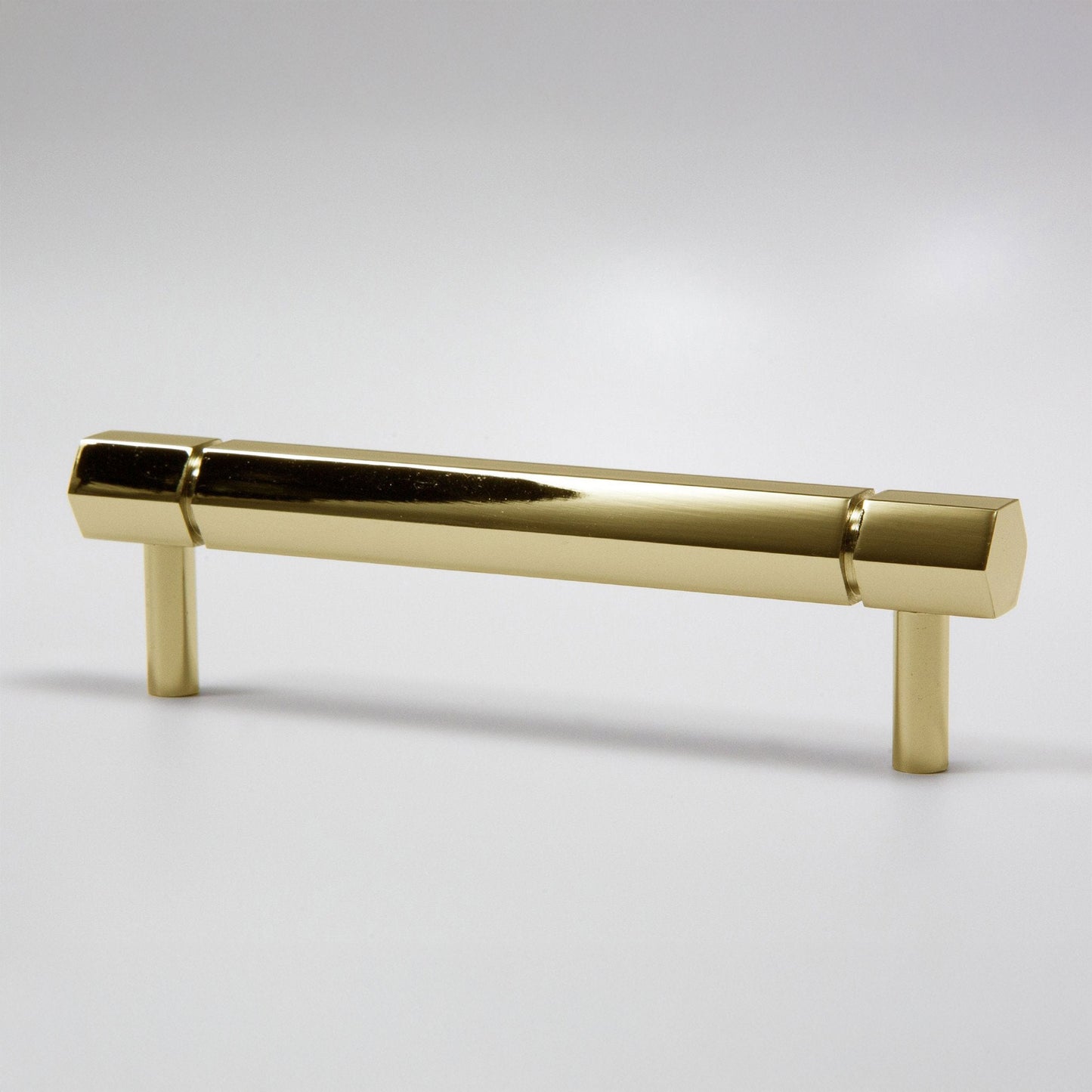 A modern hexagonal cabinet handle in a polished natural brass finish, featuring a long horizontal bar supported by two short cylindrical posts, shown on a light gray background.