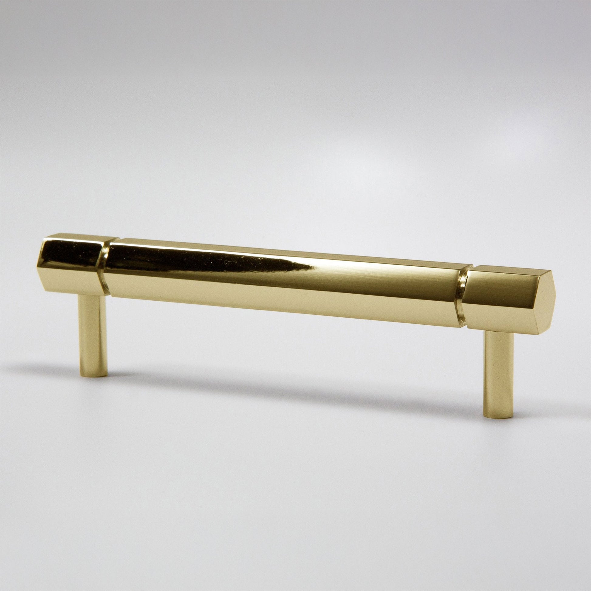 A modern hexagonal cabinet handle in a polished natural brass finish, featuring a long horizontal bar supported by two short cylindrical posts, shown on a light gray background.