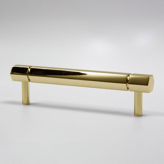 A modern hexagonal cabinet handle in a polished natural brass finish, featuring a long horizontal bar supported by two short cylindrical posts, shown on a light gray background.