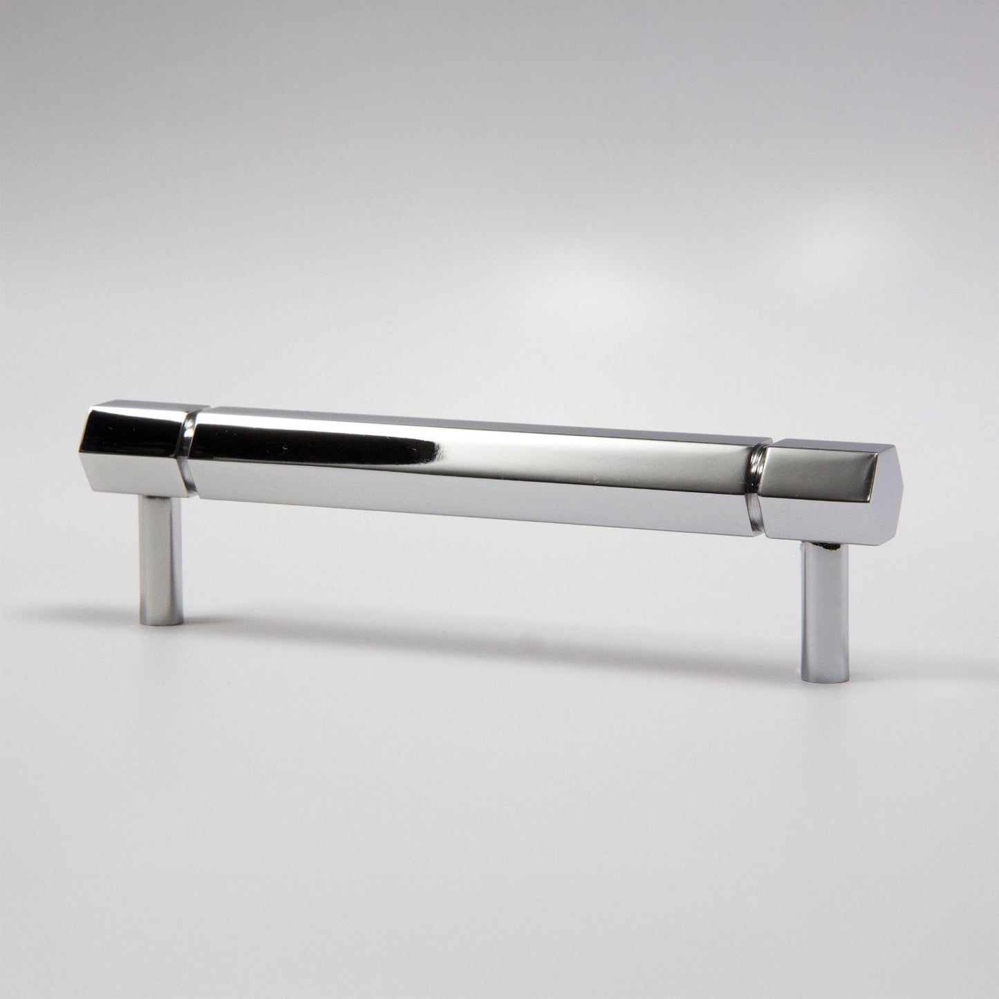 A modern hexagonal cabinet handle in polished chrome, featuring a long horizontal bar supported by two short cylindrical posts, shown on a light gray background.