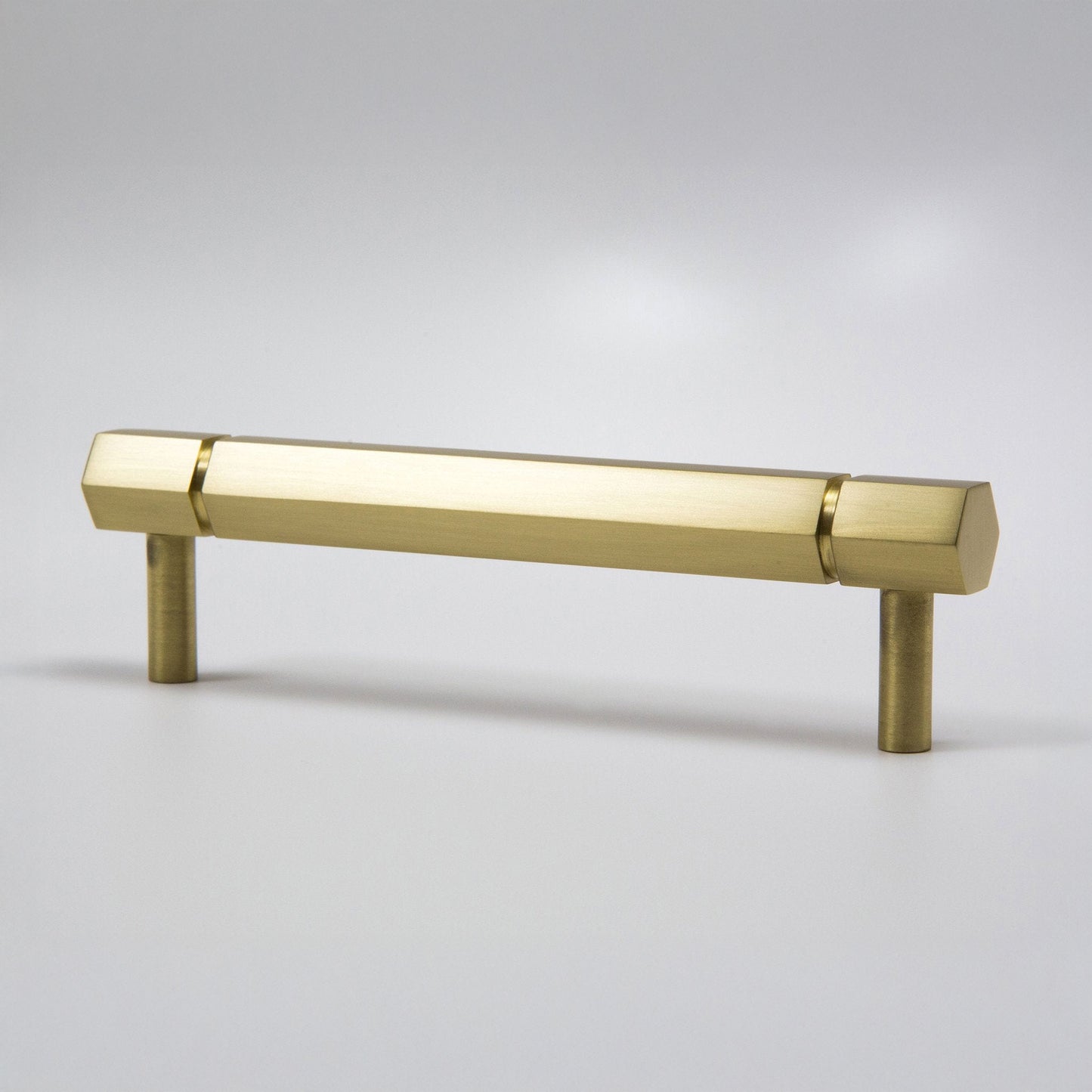 A modern hexagonal cabinet handle in satin brass, featuring a long horizontal bar supported by two short cylindrical posts, shown on a light gray background.