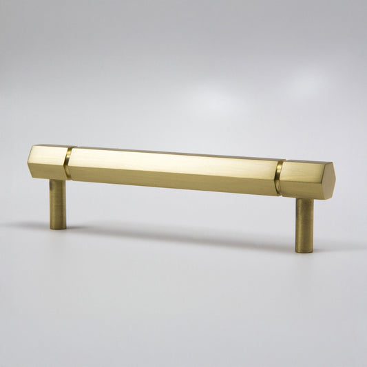 A modern hexagonal cabinet handle in satin brass, featuring a long horizontal bar supported by two short cylindrical posts, shown on a light gray background.