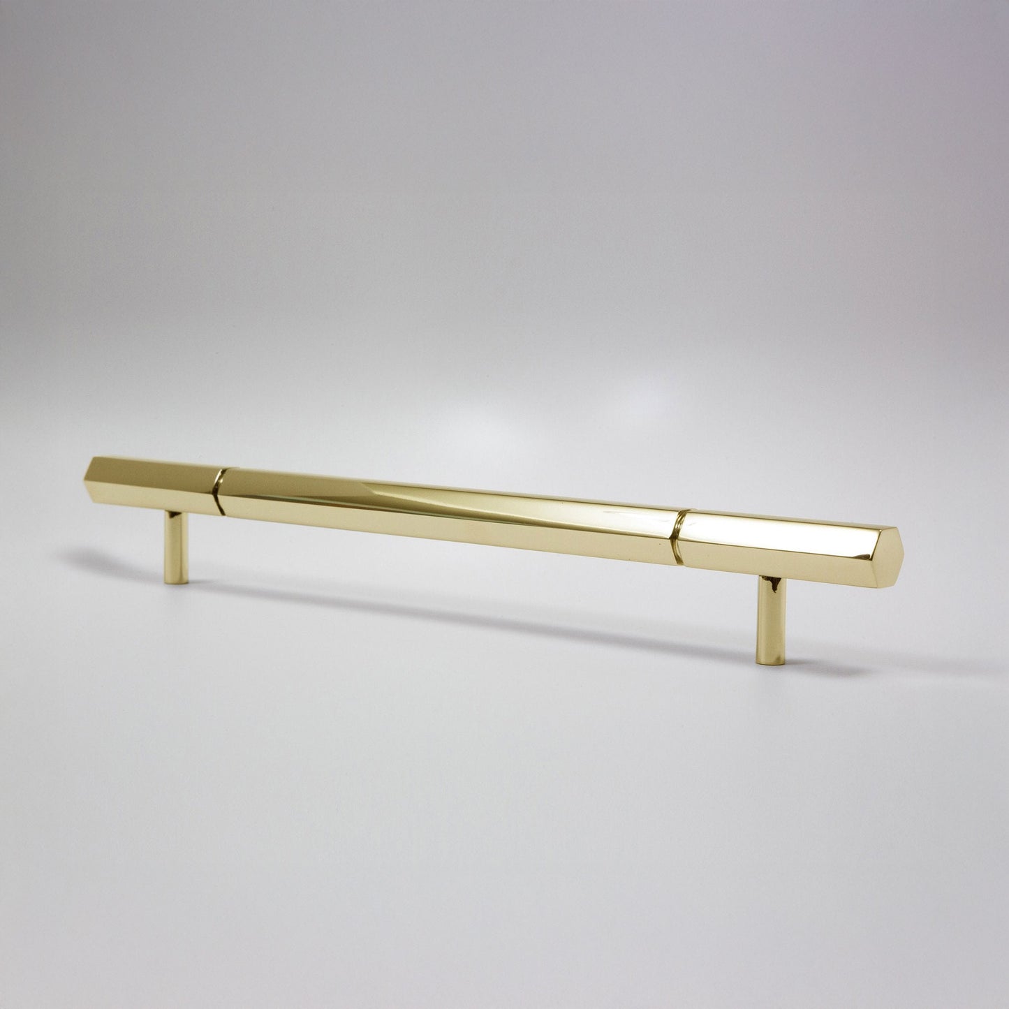 A modern hexagonal cabinet handle in polished unlaquered brass, featuring a long horizontal bar supported by two short cylindrical posts, shown on a light gray background.