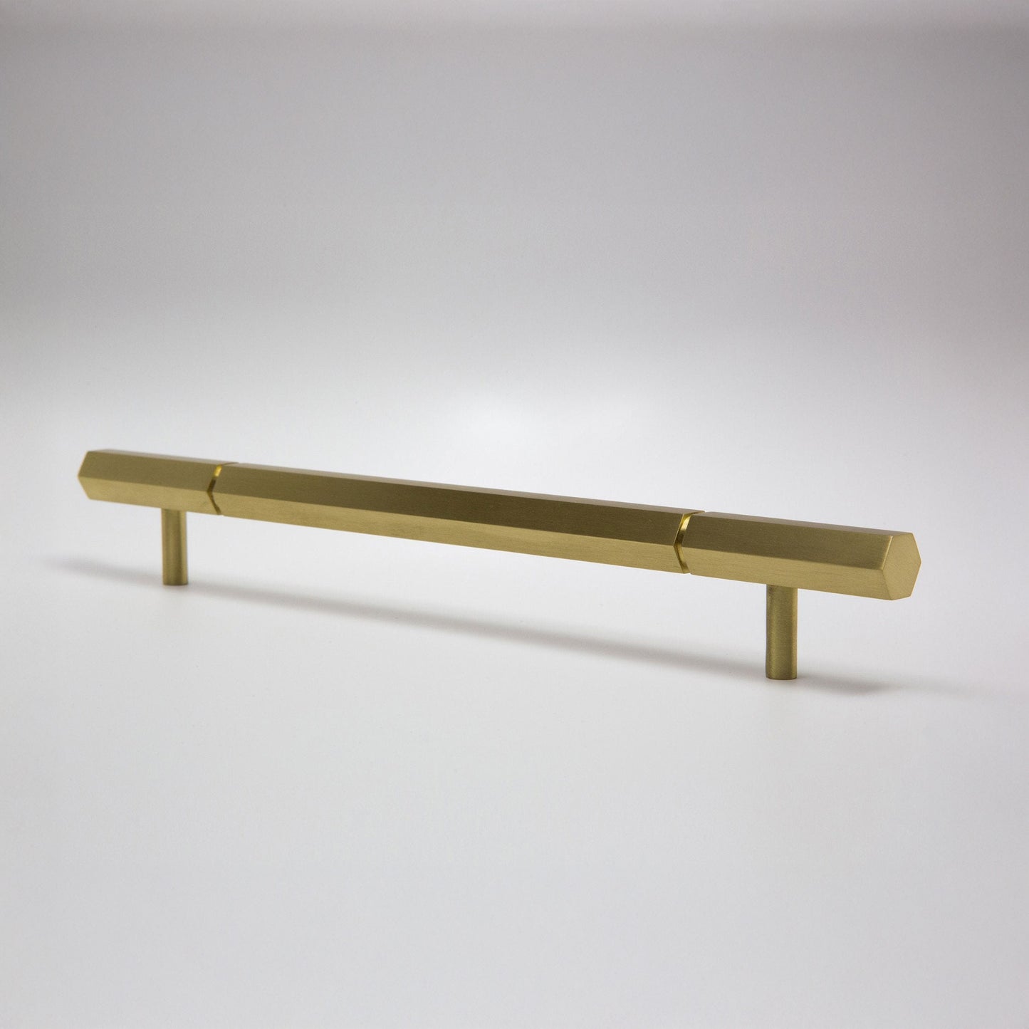 A modern hexagonal cabinet handle in satin brass, featuring a long horizontal bar supported by two short cylindrical posts, shown on a light gray background.