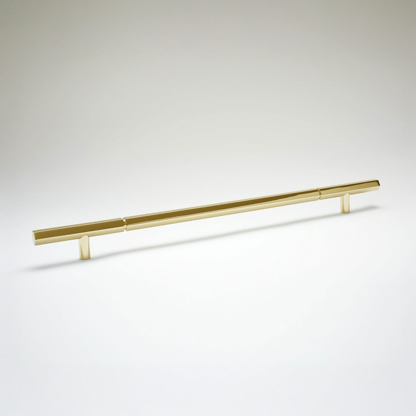 A modern hexagonal cabinet handle in unlaquered brass, featuring an extra long horizontal bar supported by two short cylindrical posts, shown on a light gray background.