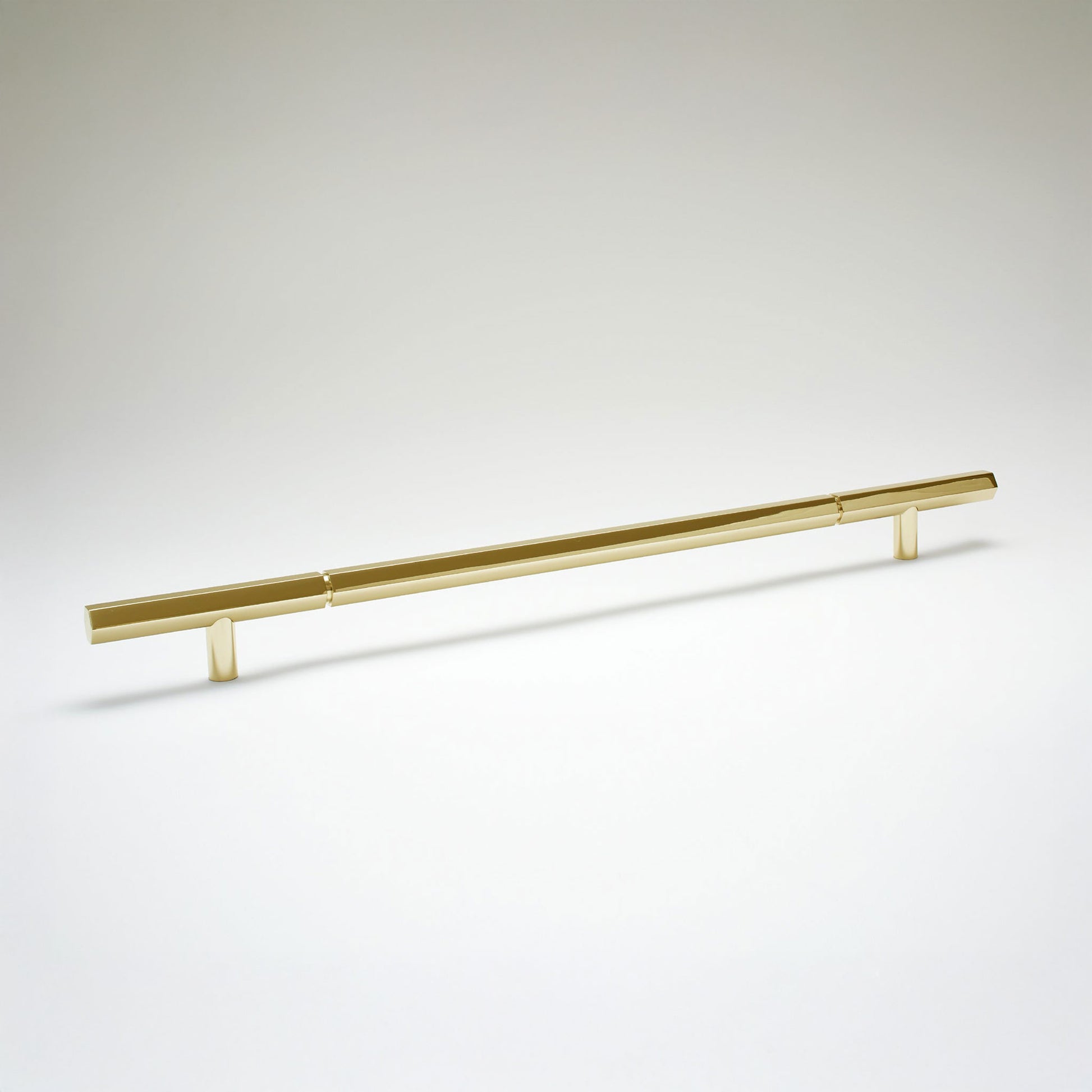 A modern hexagonal cabinet handle in unlaquered brass, featuring an extra long horizontal bar supported by two short cylindrical posts, shown on a light gray background.