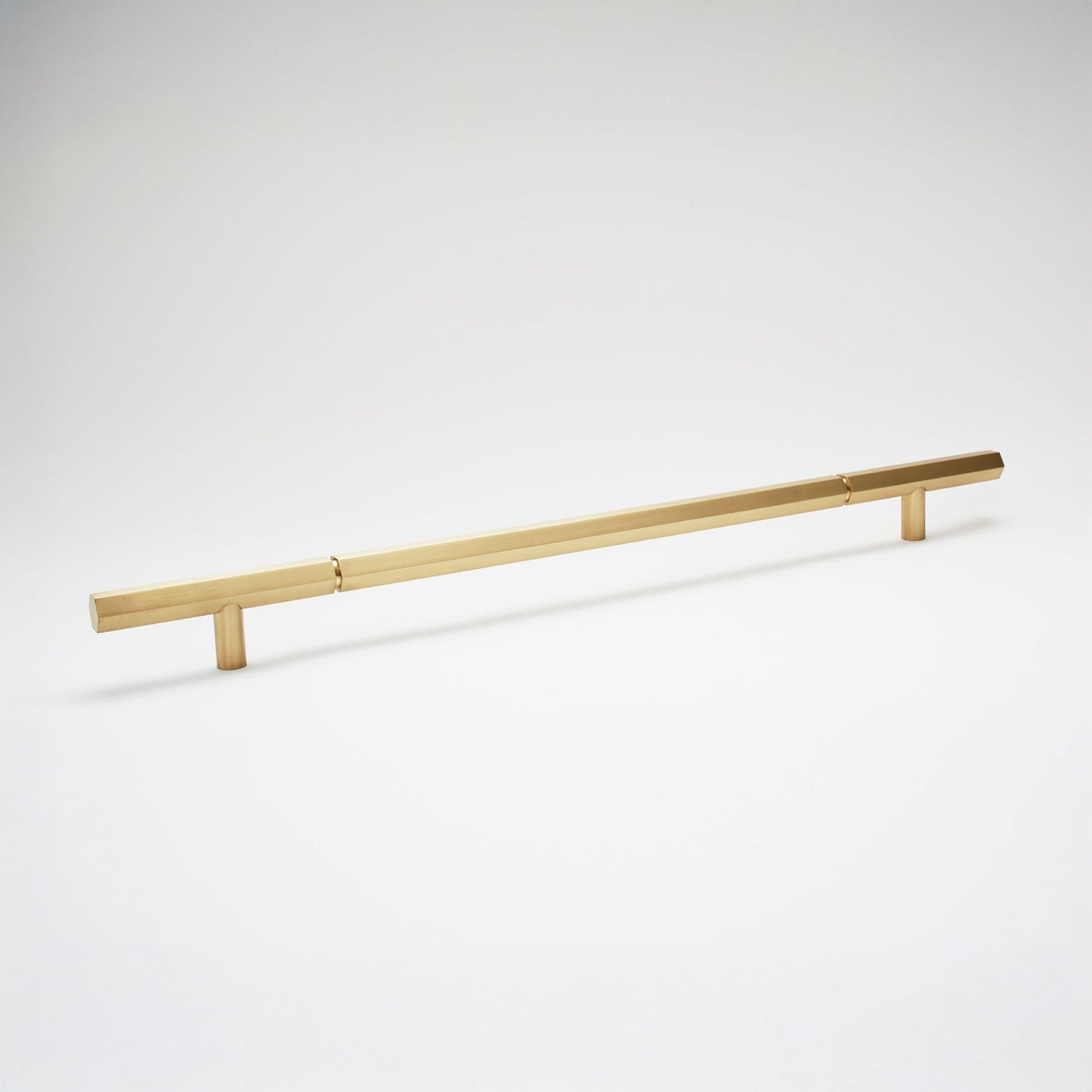 A modern hexagonal cabinet handle in satin brass, featuring an extra long horizontal bar supported by two short cylindrical posts, shown on a light gray background.