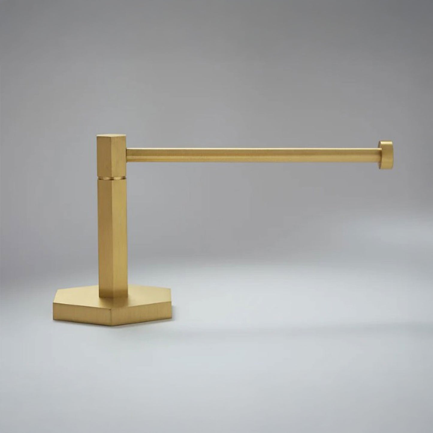 A modern wall-mounted paper holderr with a polished brass finish, featuring a square post and a flat base, shown against a neutral gray background.