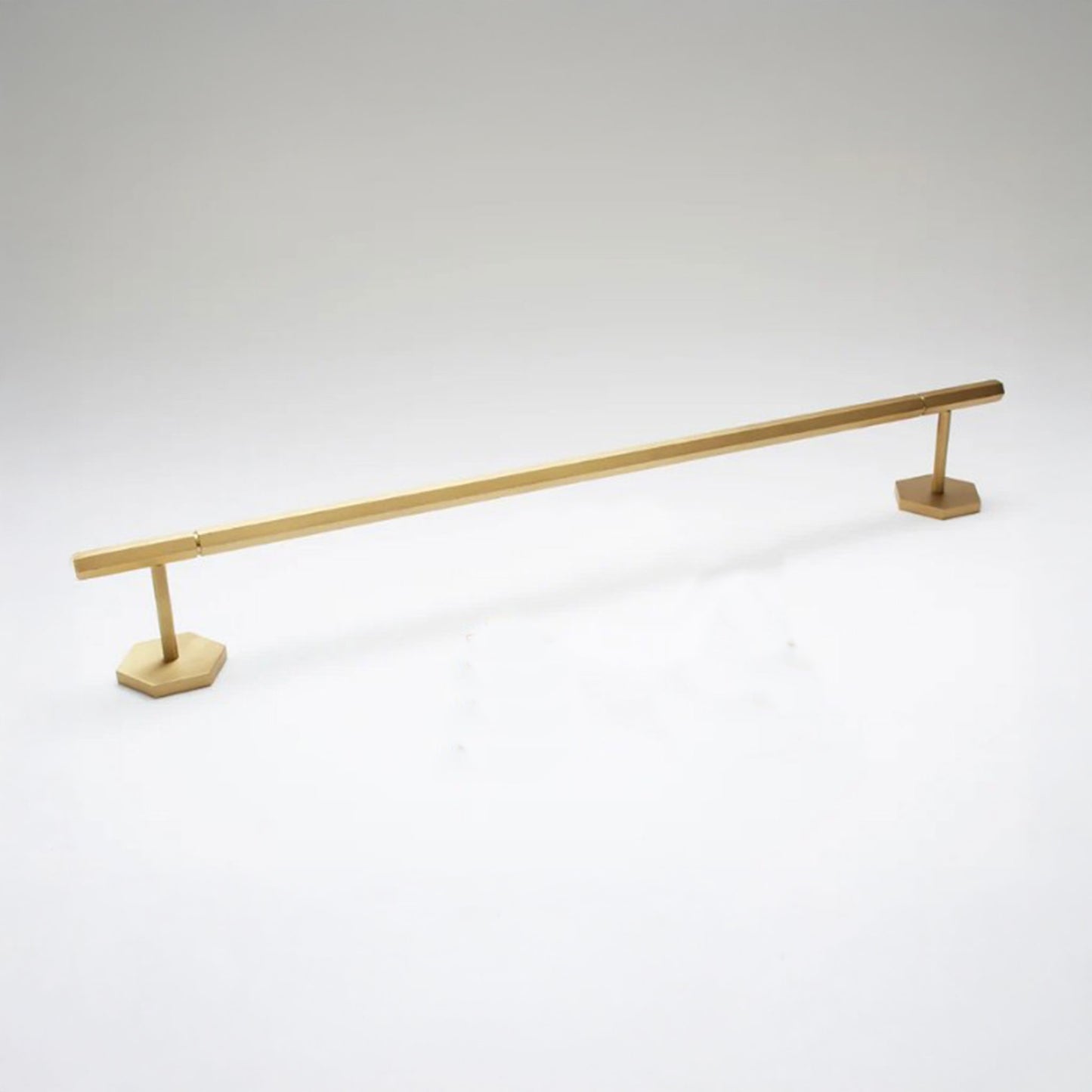 A modern brass towel bar with a sleek, minimalist design, featuring a long horizontal bar supported by two hexagonal bases, shown on a light gray background.