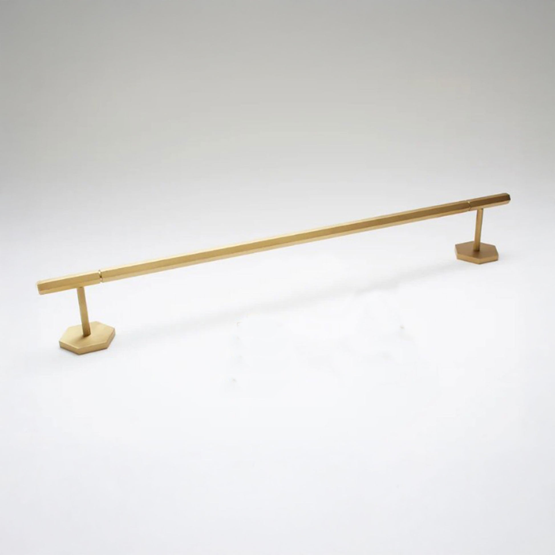 A modern brass towel bar with a sleek, minimalist design, featuring a long horizontal bar supported by two hexagonal bases, shown on a light gray background.