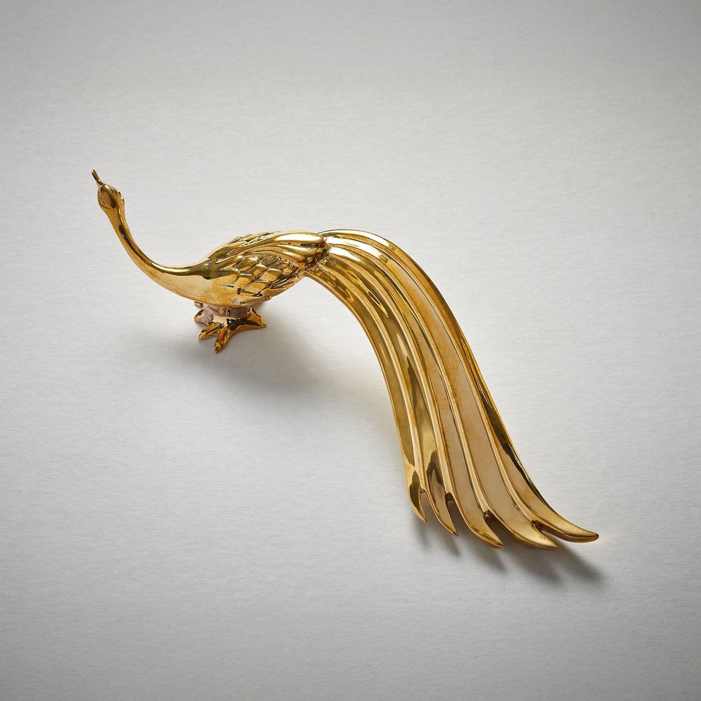 A polished unlaquered brass metal cabinet pull handle shaped like a stylized peacock with a long flowing tail, photographed on a plain light gray background.