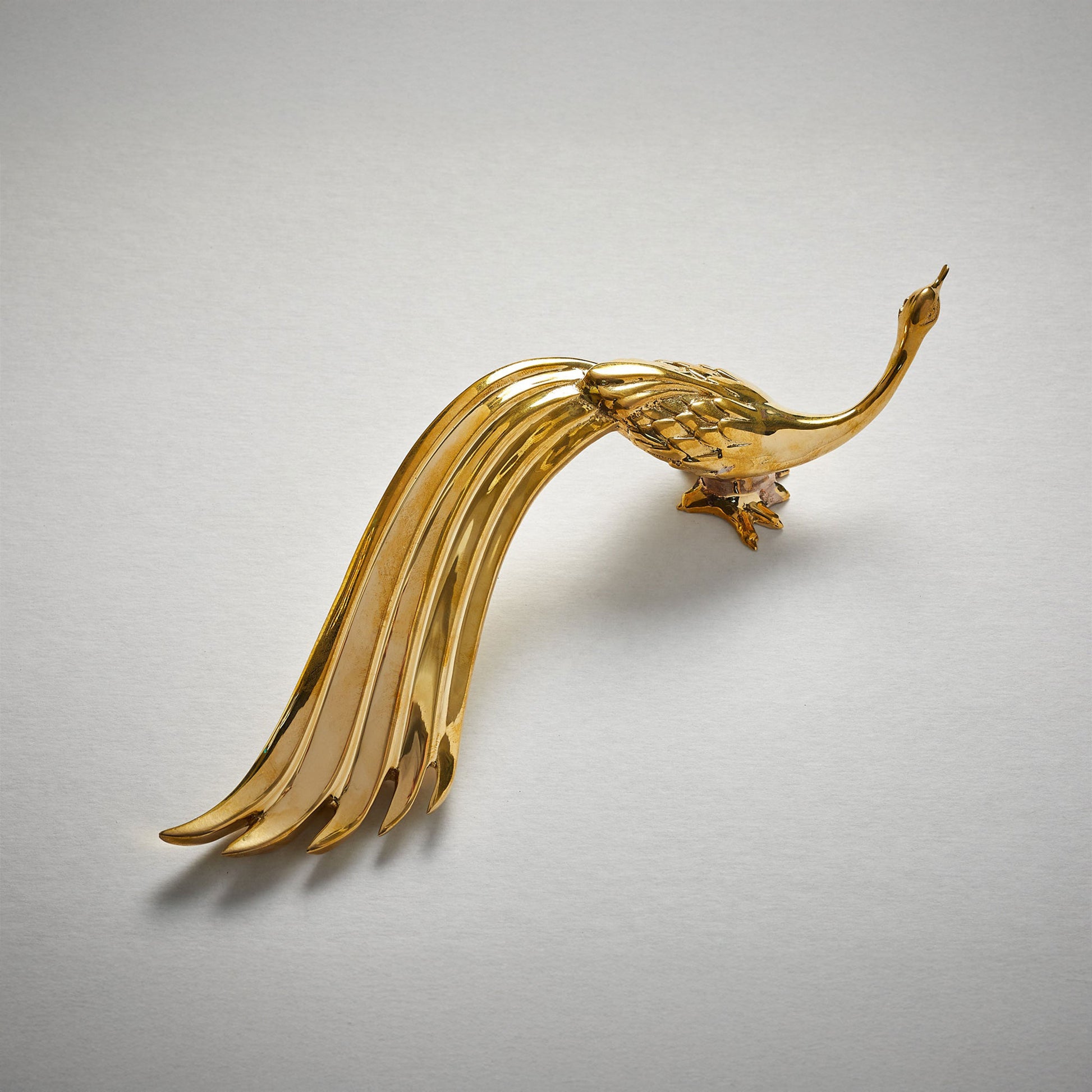 A polished unlaquered brass metal cabinet pull handle shaped like a stylized peacock with a long flowing tail, photographed on a plain light gray background.