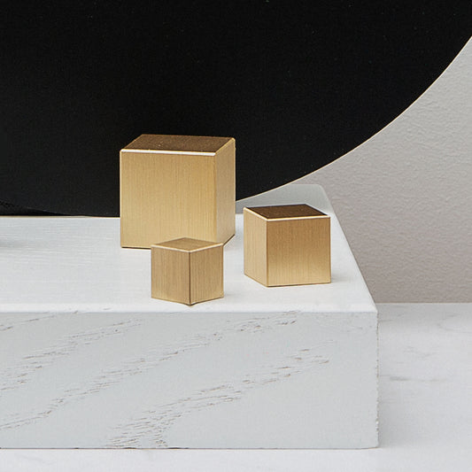 Three gold cube-shaped cabinet knobs on a marble surface with a black and gray background