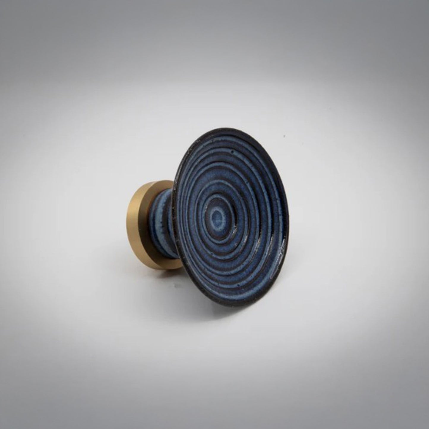 A round ceramic cabinet knob or pull with a dark blue concentric ring patterned top and a gold-toned cylindrical base, photographed on a light gray background.