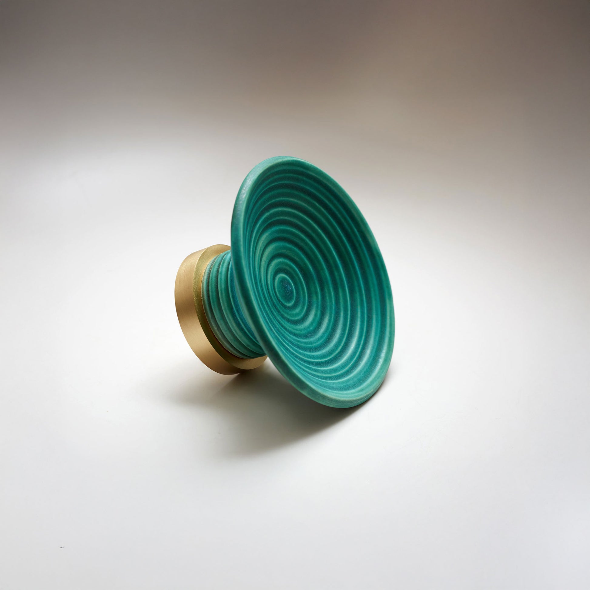 A round ceramic cabinet knob or pull with a teal concentric ring patterned top and a gold-toned cylindrical base, photographed on a light gray background.