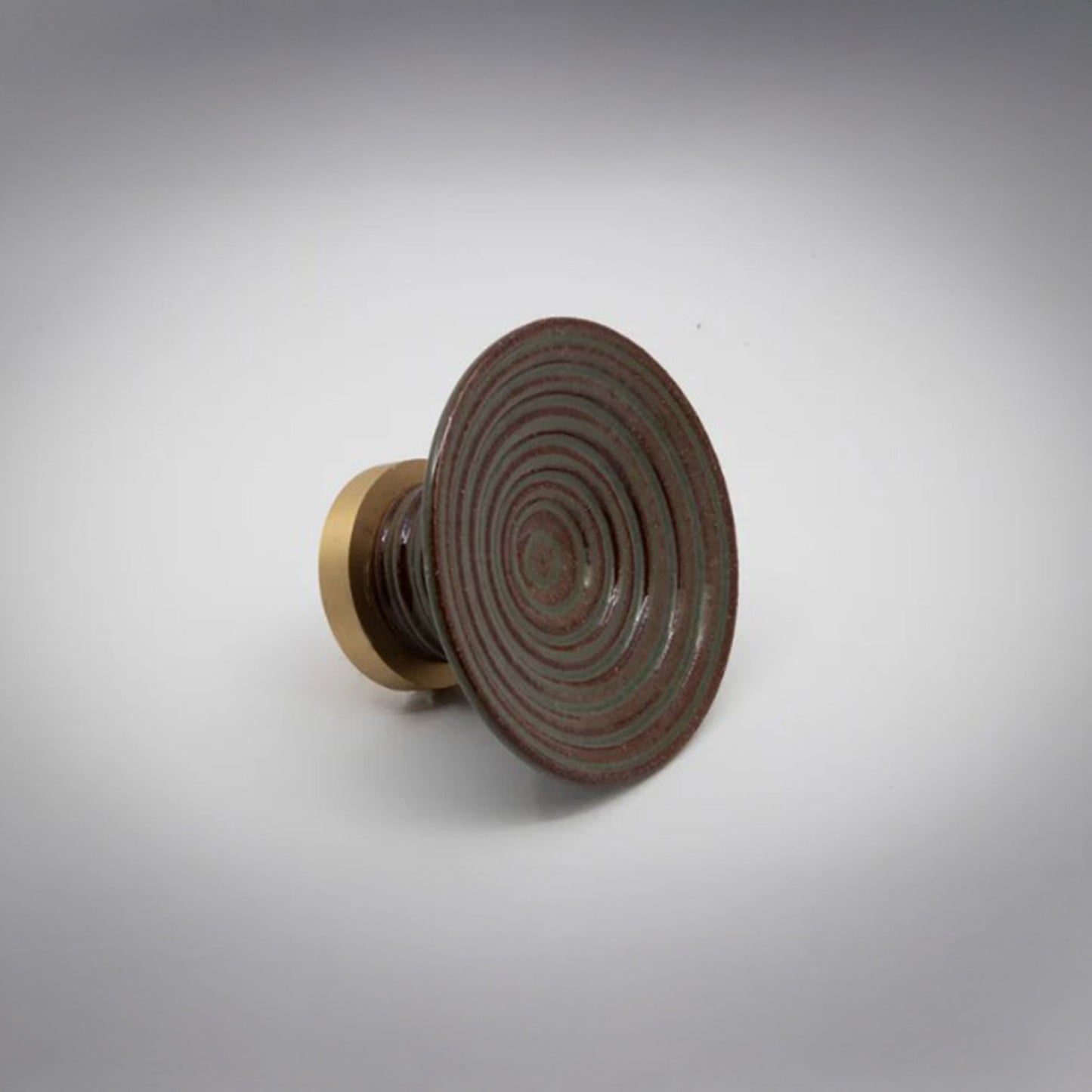 A round ceramic cabinet knob or pull with a sage green and terra cotta concentric ring patterned top and a gold-toned cylindrical base, photographed on a light gray background.