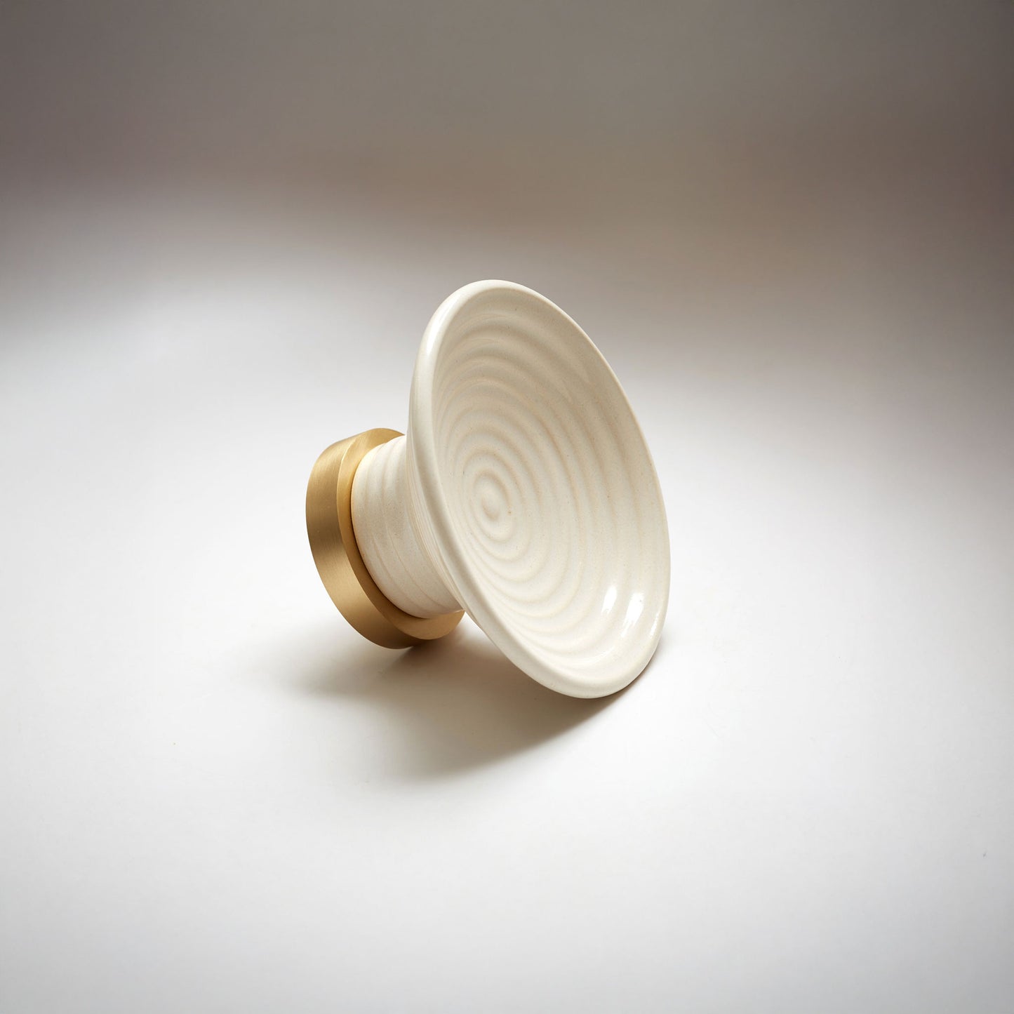 A round ceramic cabinet knob or pull with a soft white concentric ring patterned top and a gold-toned cylindrical base, photographed on a light gray background.