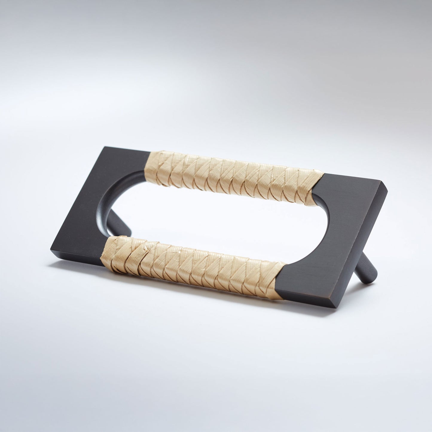 A modern dark rubbed bronze metal cabinet pull handle with an oval cutout, wrapped with a light colored natural beige woven leather,  shown on a white background.