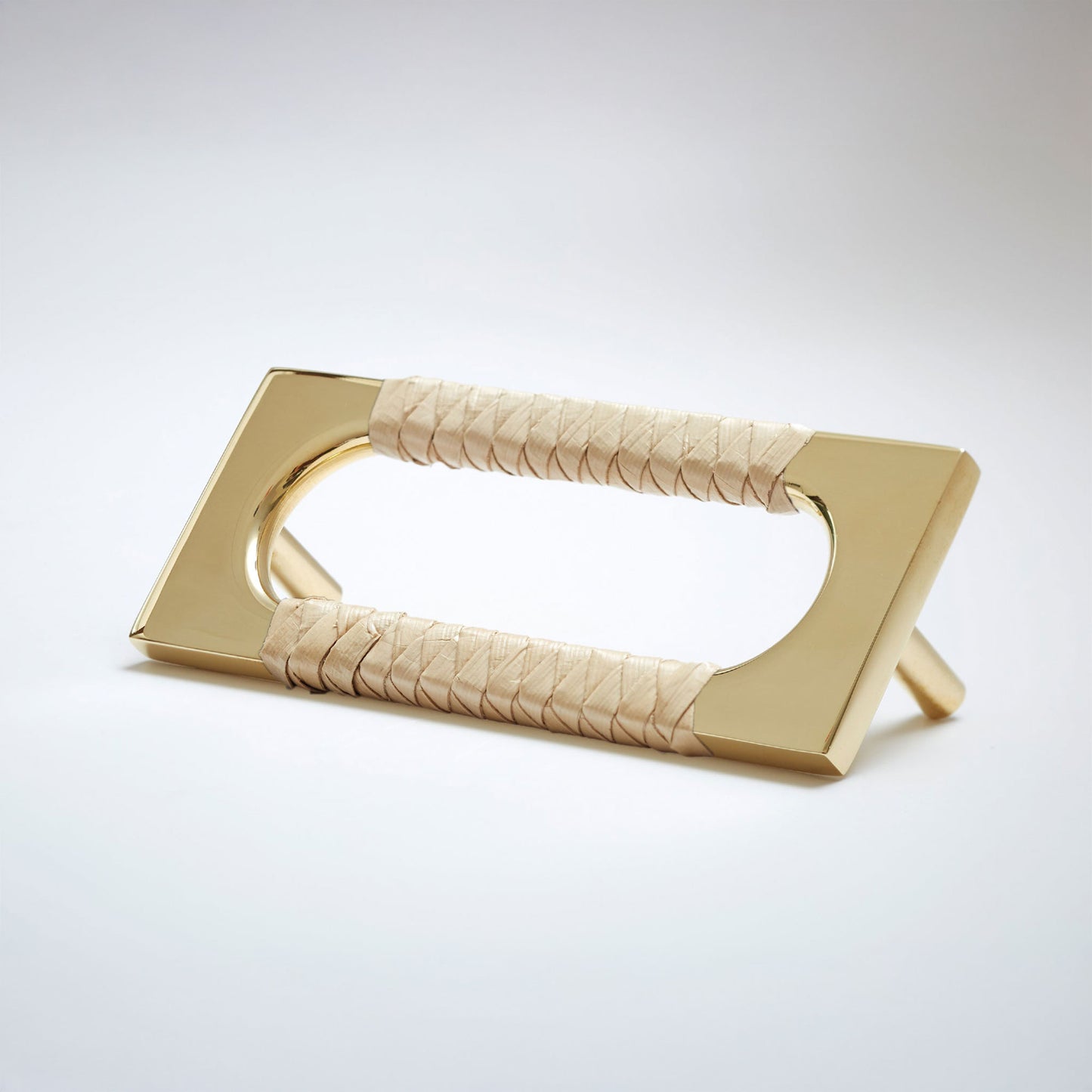 A modern polished natural brass metal cabinet pull handle with an oval cutout, wrapped with a light colored natural beige screwpine leaf,  shown on a white background.