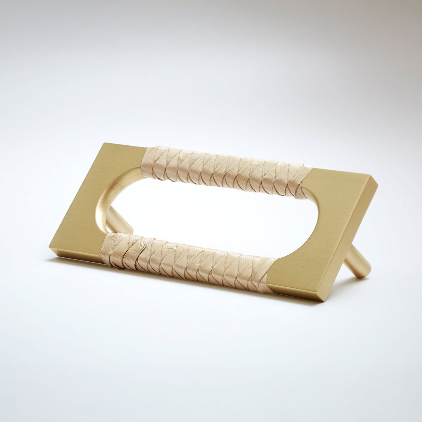 A modern satin brass metal cabinet pull handle with an oval cutout, wrapped with a light colored natural beige screwpine leaf,  shown on a white background.