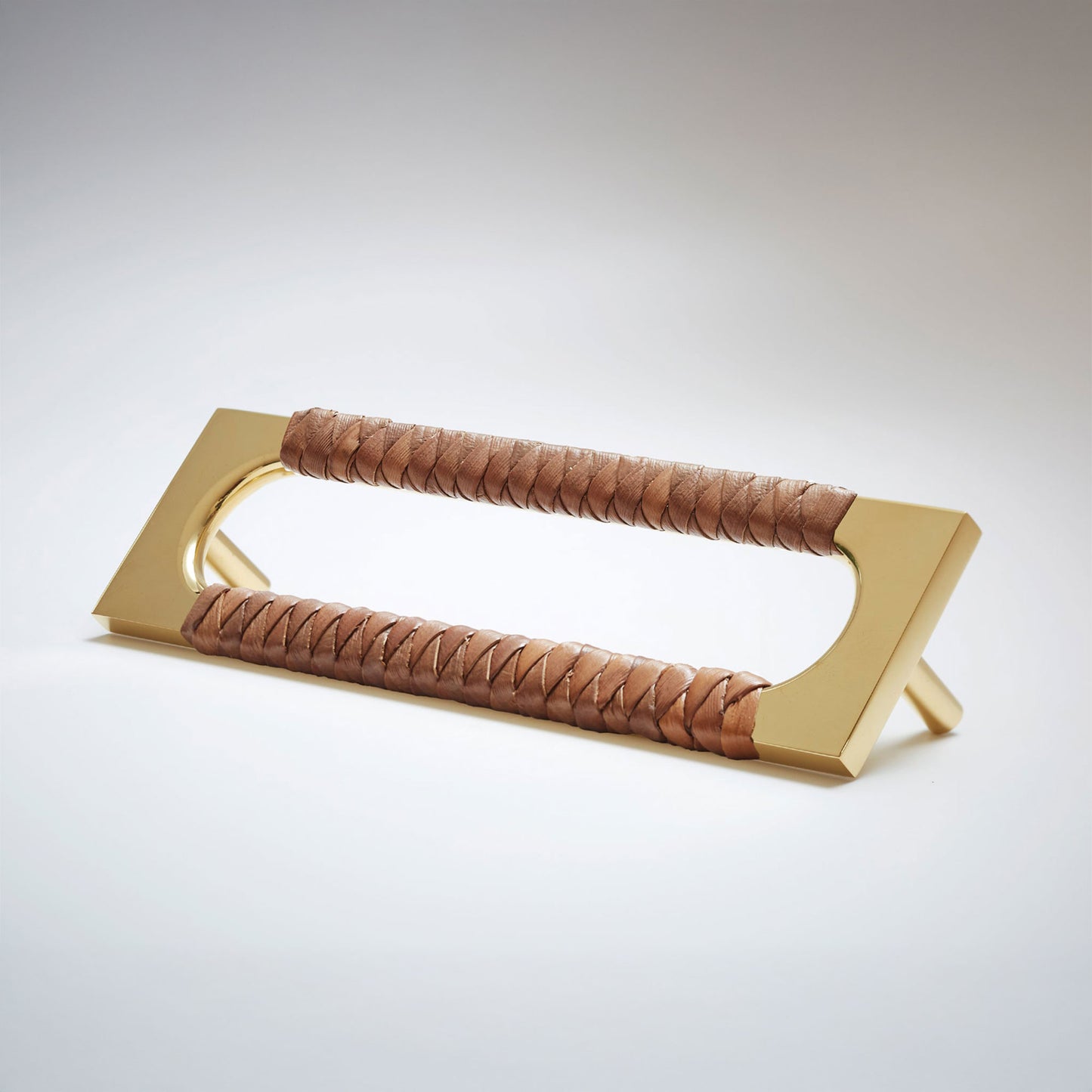 A modern brass door pull handle featuring a curved, open-top design with two wrapped brown leather handles, shown on a neutral light gray background.
