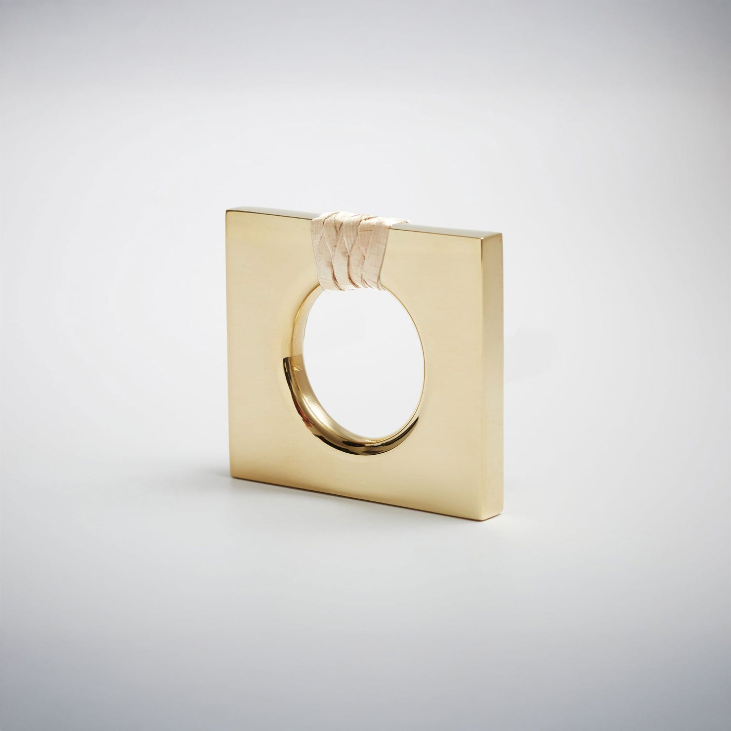 A petite square-shaped polished natural brass cabinet knob with a circular cutout, featuring a light natural woven cord wrapped around the top edge, set against a white background. 