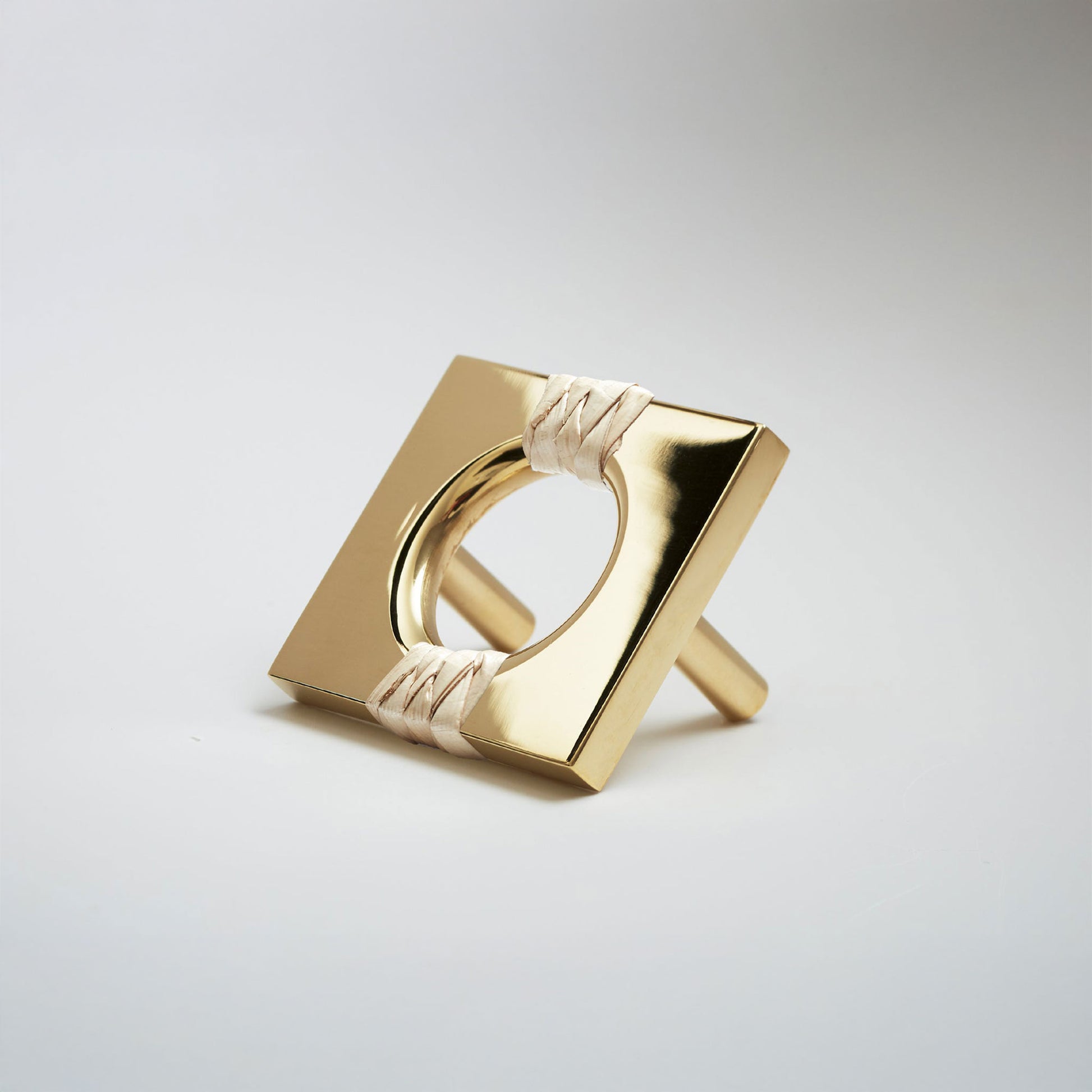 A shiny brass metal geometric cabinet pull with a square frame and circular center, wrapped with a light natural leather at the corners, shown on a white background.