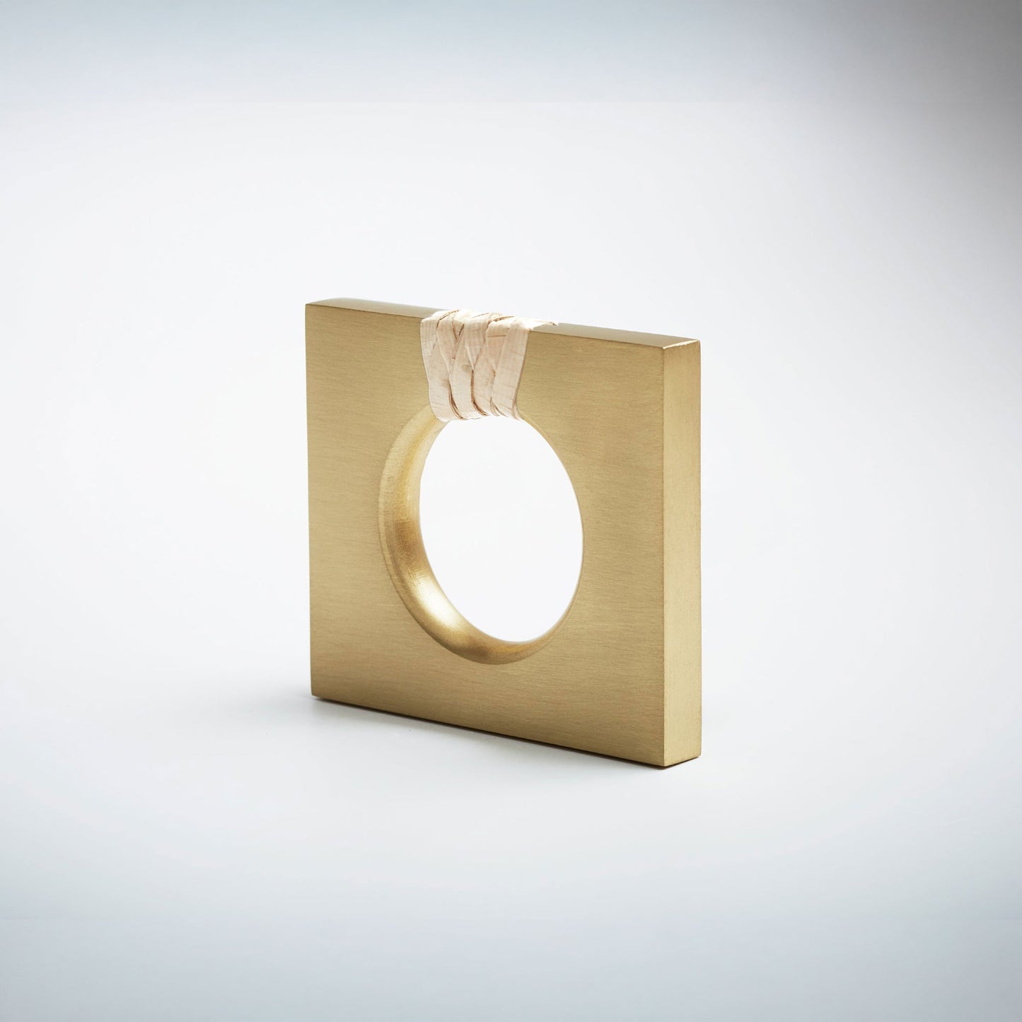 A petite square-shaped satin brass cabinet knob with a circular cutout, featuring a light natural woven cord wrapped around the top edge, set against a white background. 