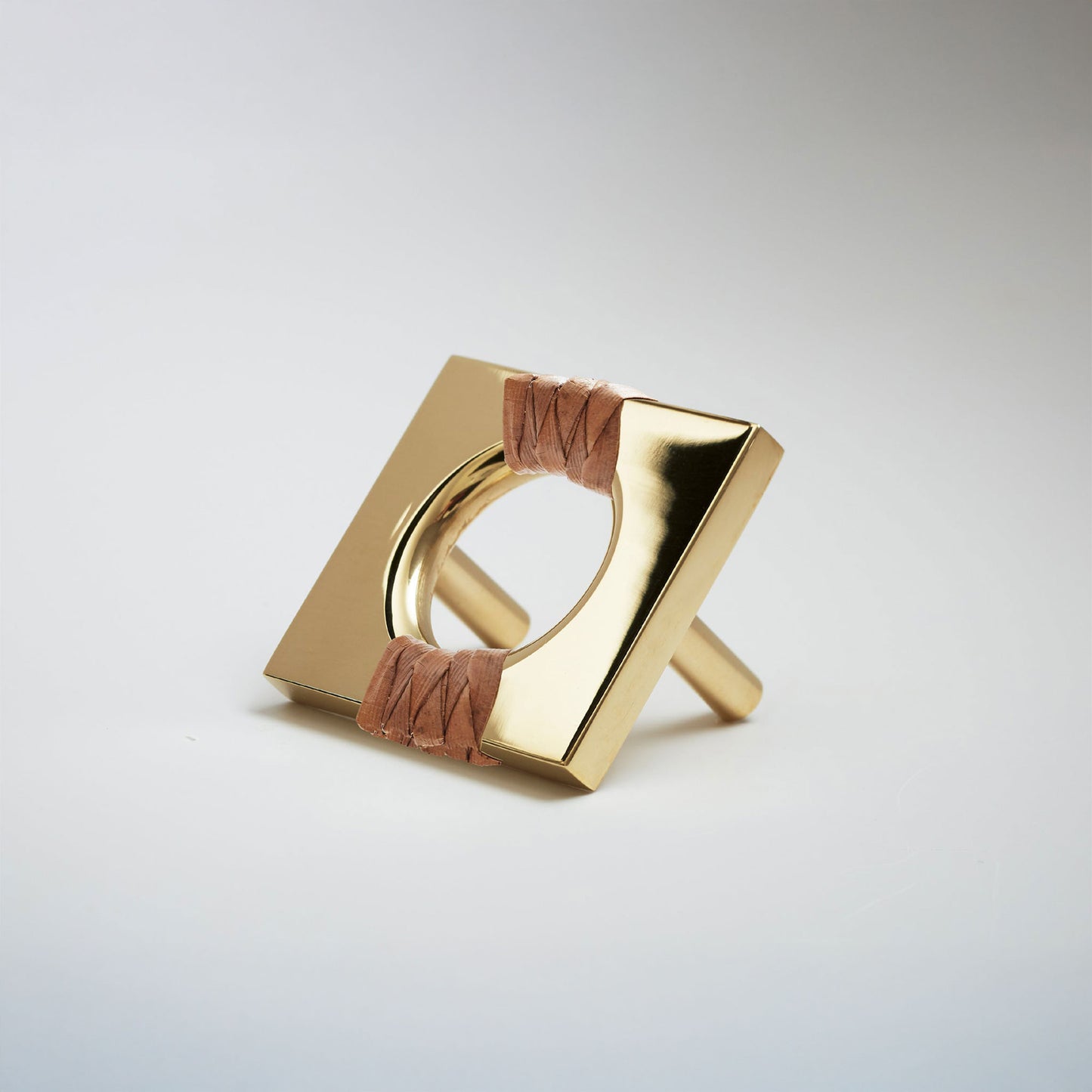 A shiny brass geometric cabinet pull with a square frame and circular center, wrapped with brown pine reed on either side, shown on a white background.