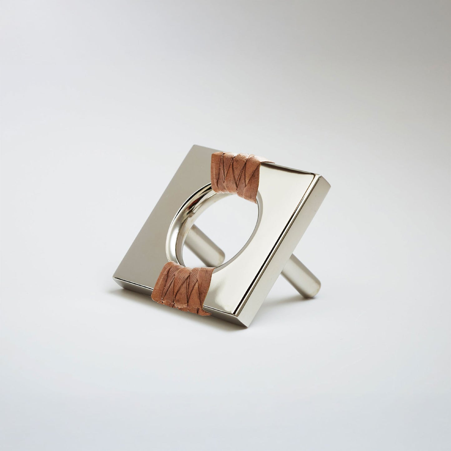 A shiny polished nickel metal geometric cabinet pull with a square frame and circular center, wrapped with brown leather at the corners, shown on a white background.