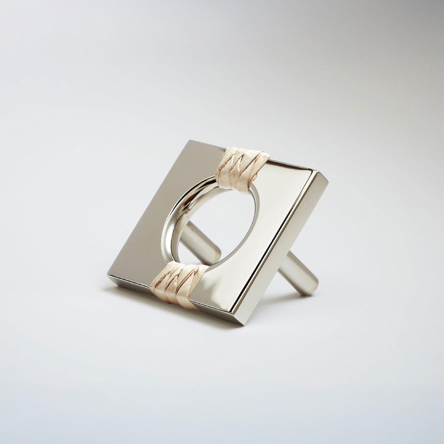 A shiny polished nickel metal geometric cabinet pull with a square frame and circular center, wrapped with a light natural leather at the corners, shown on a white background.